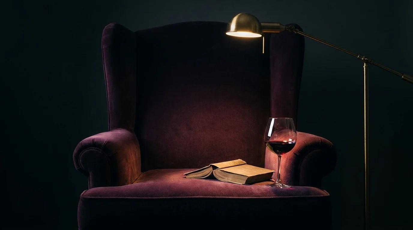 Open hardback face-down on a deep plum velvet armchair beside red wine under a brass reading lamp's warm pool of light