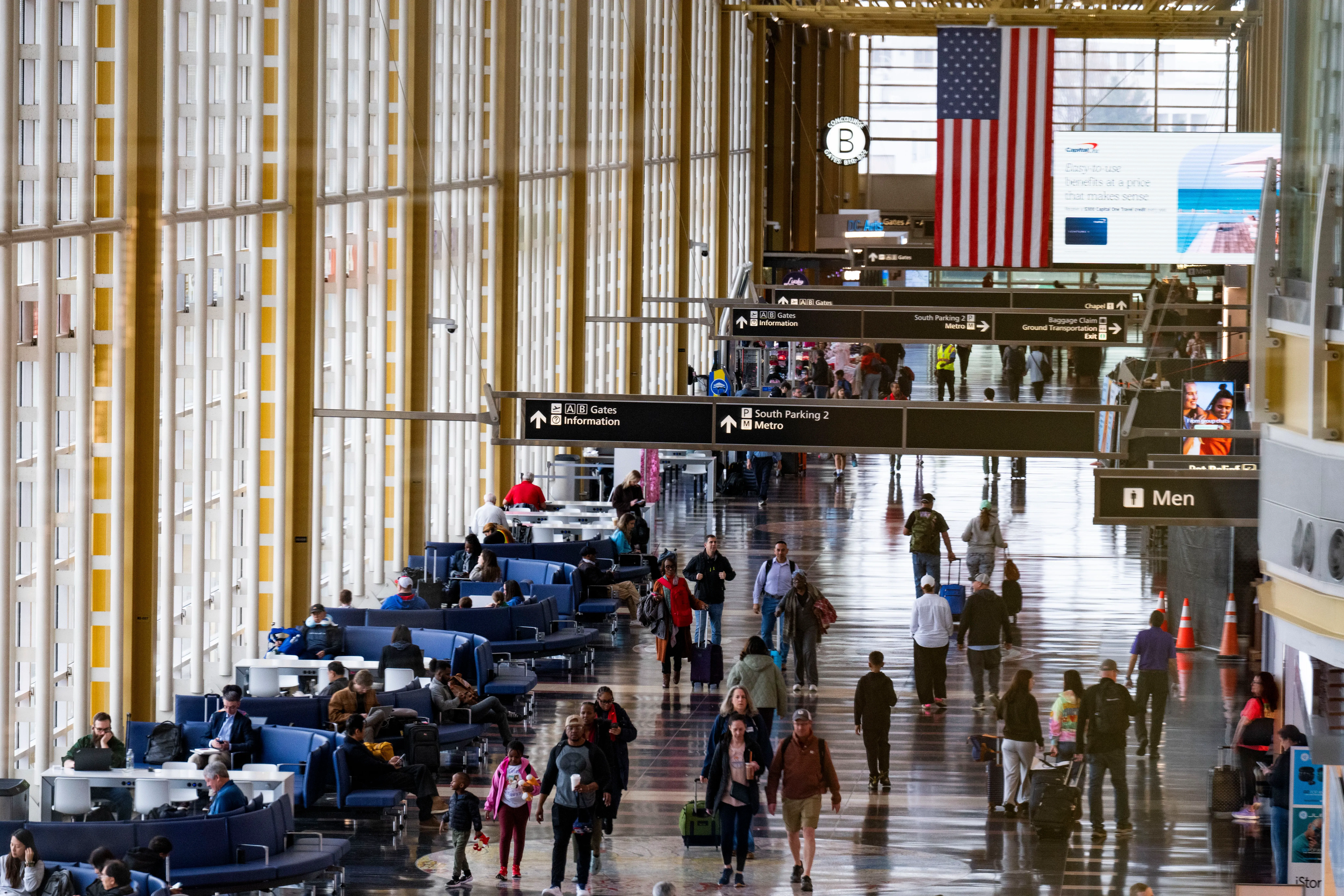 ازدحام في مطار حديث مع مسافرين يتنقلون بين المقاعد، ولافتات توجيهية، وعلم أمريكي معلق في الأعلى، مما يعكس الحركة النشطة للسفر الجوي.