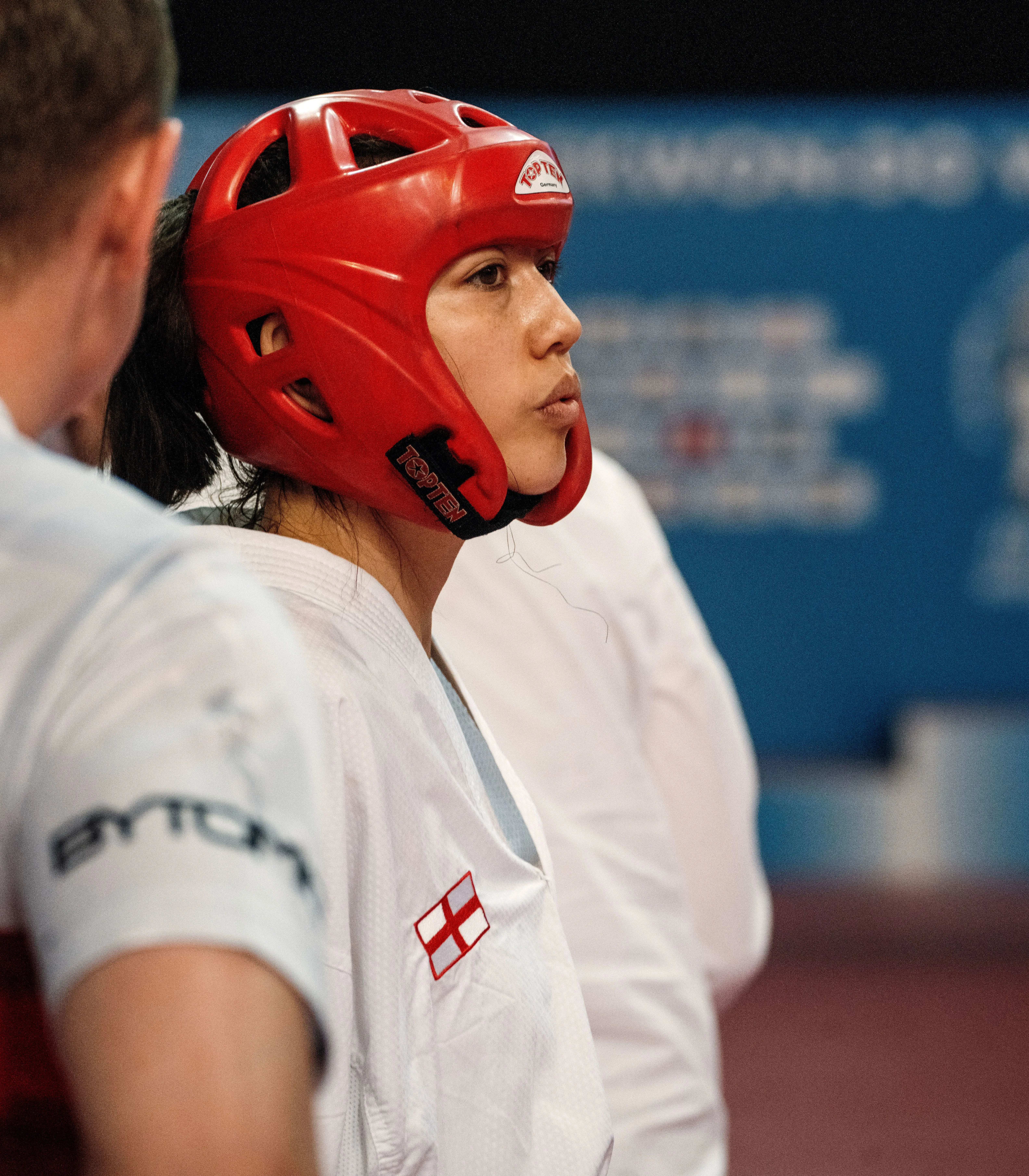 لاعبة تايكوندو ترتدي خوذة حمراء وتبدو مركّزة، بينما تستعد لمنافسة في بطولة عالمية، مع خلفية توضح أجواء البطولة.