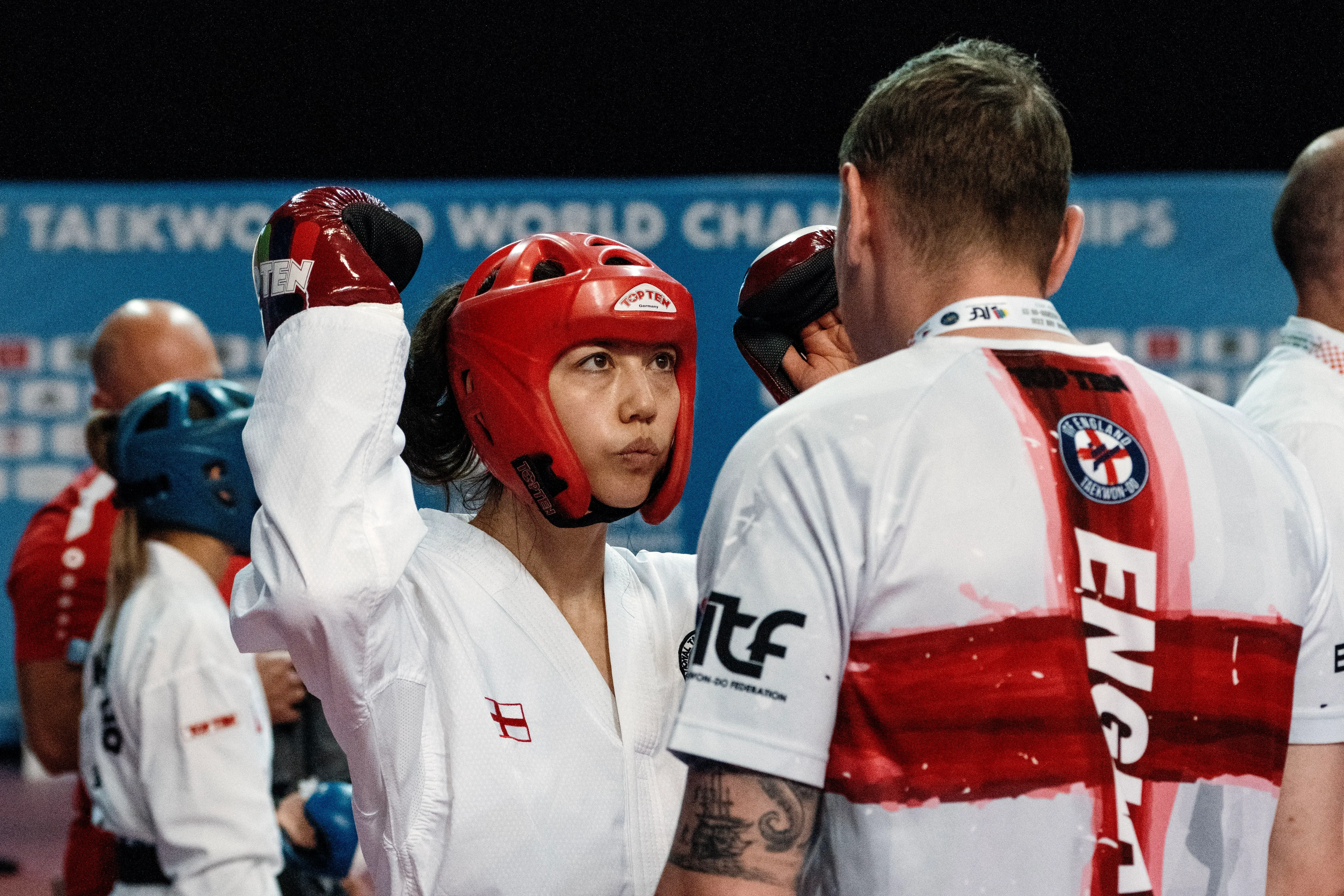 منافسة تايكوندو في بطولة العالم، حيث يرتدي اللاعب خوذة حمراء ويتلقى توجيهات من مدربه، مع خلفية توضح أجواء البطولة.