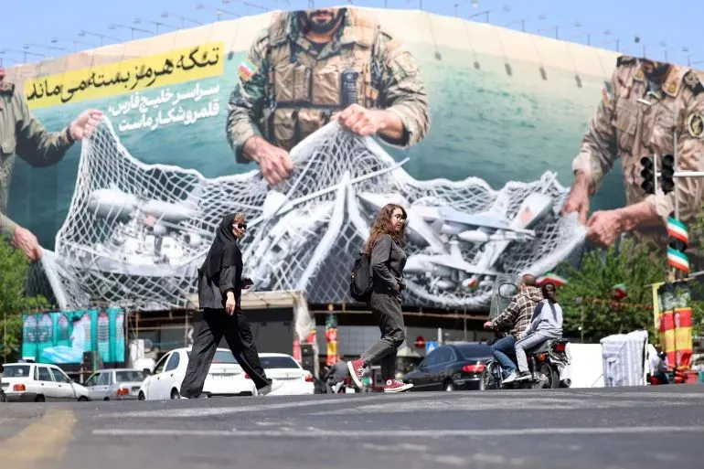 امرأتان تعبران الشارع أمام جدارية كبيرة تظهر جندياً يحمل أسلحة، تعكس التوترات العسكرية في الشرق الأوسط.