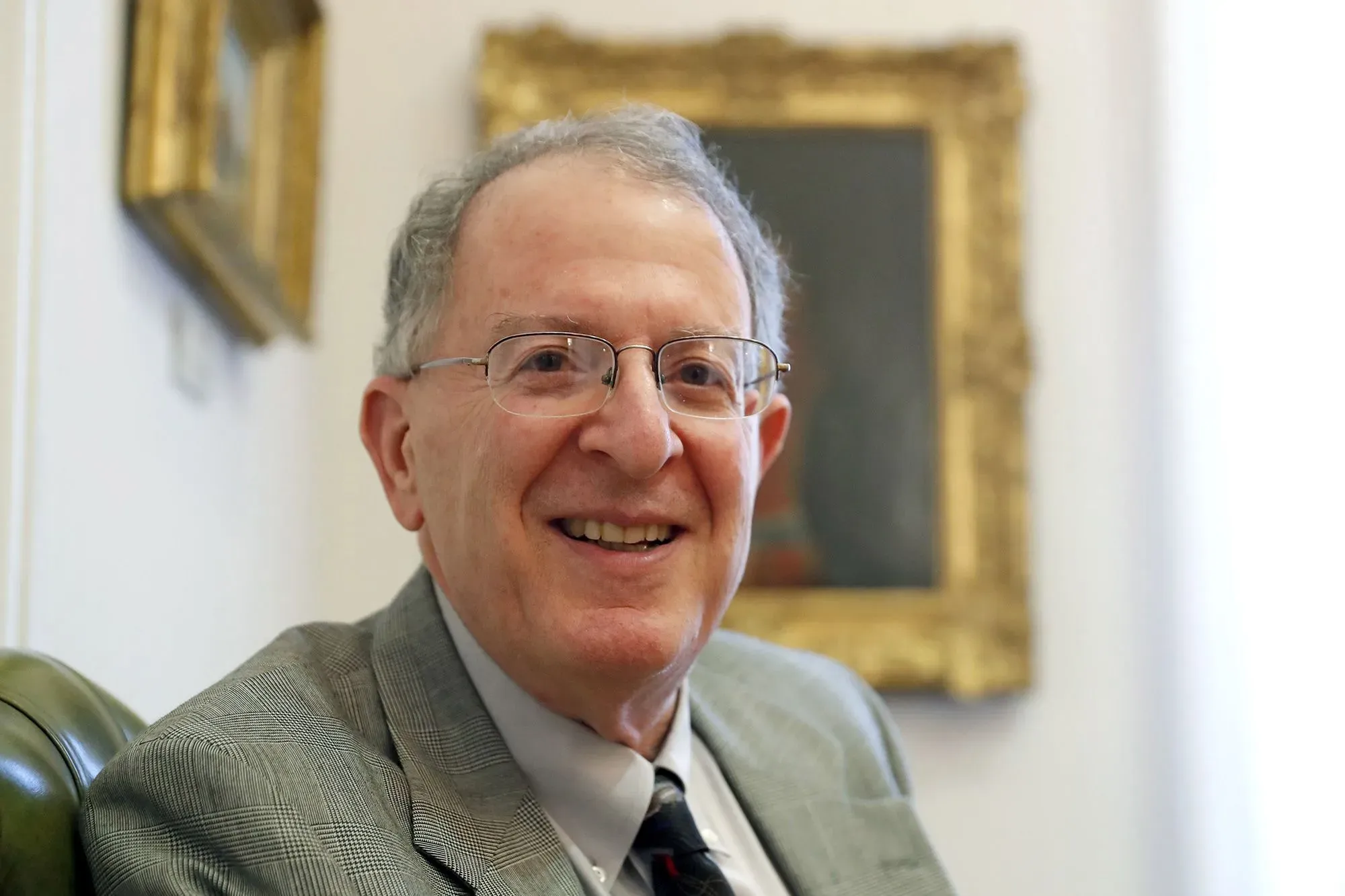 عالم مبتسم يرتدي نظارات، يجلس في مكتب مع إطار صورة خلفه، يعكس إنجازاته في البحث العلمي.