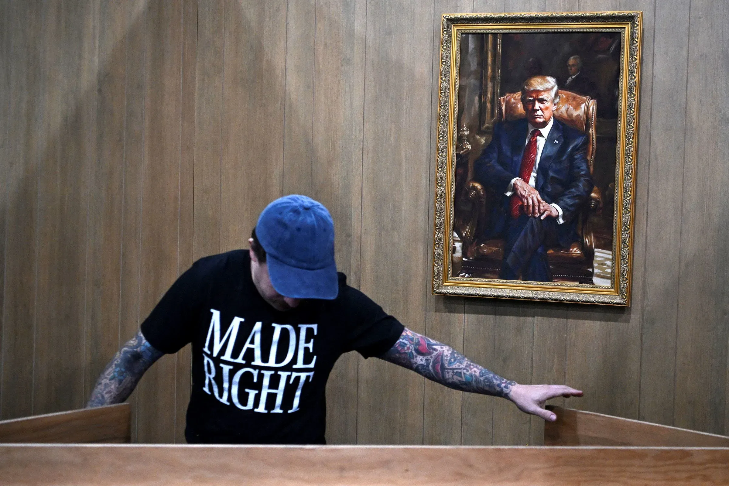 شخص يرتدي قميصًا مكتوبًا عليه "MADE RIGHT" يقف أمام لوحة للرئيس السابق دونالد ترامب، في سياق مؤتمر العمل السياسي المحافظ.