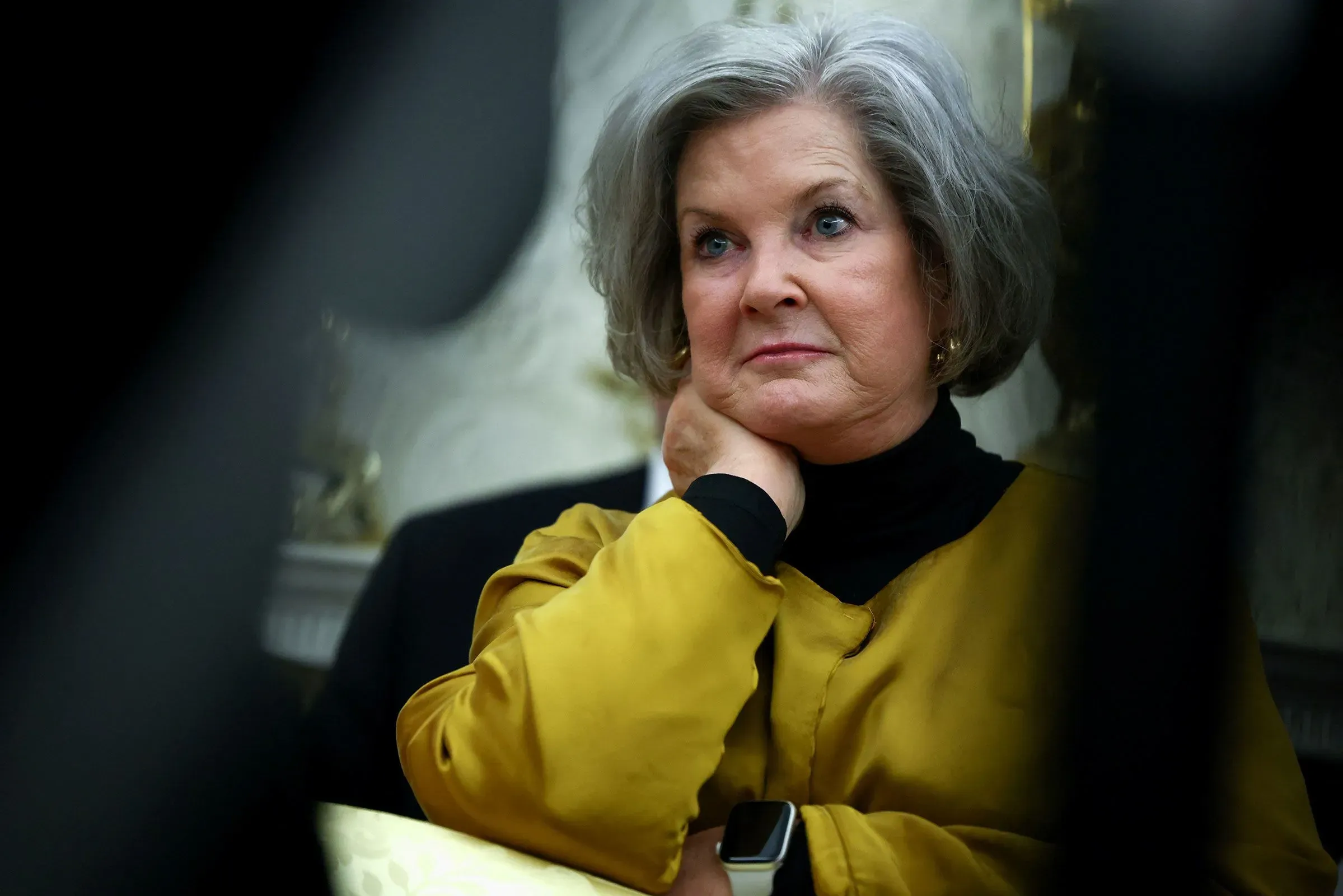 امرأة ترتدي سترة صفراء، تبدو عميقة التفكير أثناء وجودها في غرفة رسمية. تعكس تعابير وجهها مشاعر الجدية والتأمل، مما يتماشى مع موضوع المقابلة السياسية.