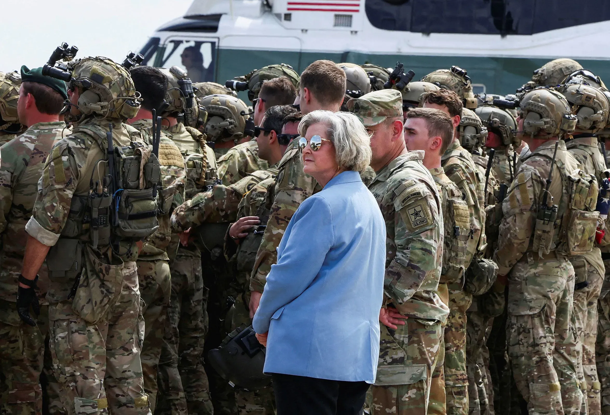 امرأة ترتدي سترة زرقاء تقف بين مجموعة من الجنود العسكريين، مع طائرة هليكوبتر في الخلفية، تعكس أجواء رسمية.