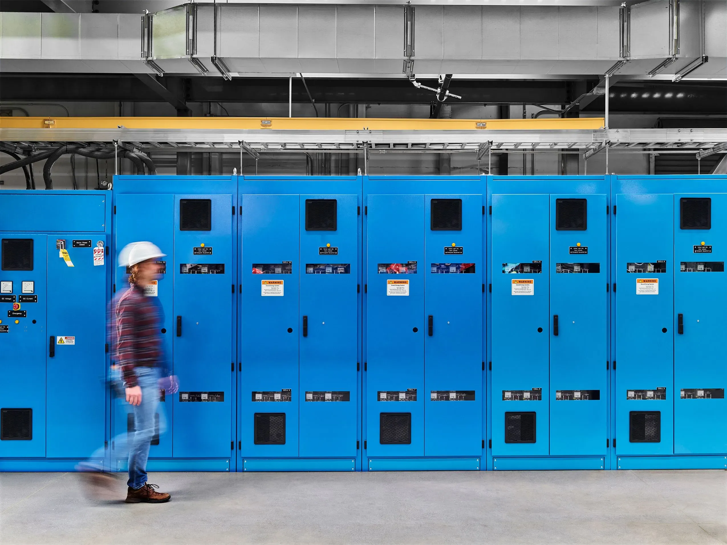 رجل يرتدي خوذة يسير بجانب صف من الخزانات الزرقاء في منشأة طاقة الاندماج النووي، مما يعكس تقدم التكنولوجيا في هذا المجال.