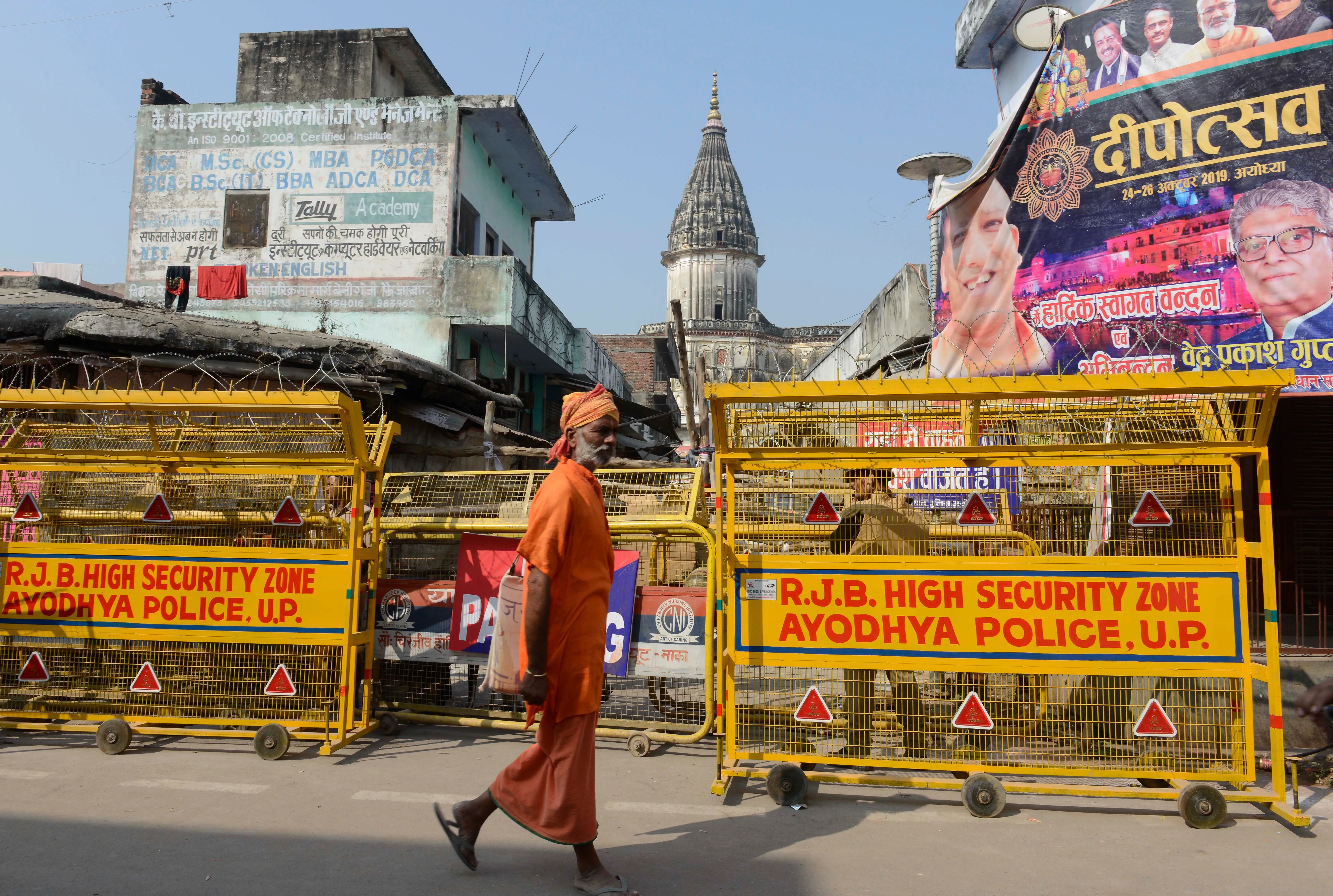 صورة لرجل يرتدي ملابس تقليدية يسير بجانب حواجز أمنية في منطقة أيوديا، خلفه معالم تاريخية وإعلانات ملونة، تعكس التوترات الثقافية والدينية.