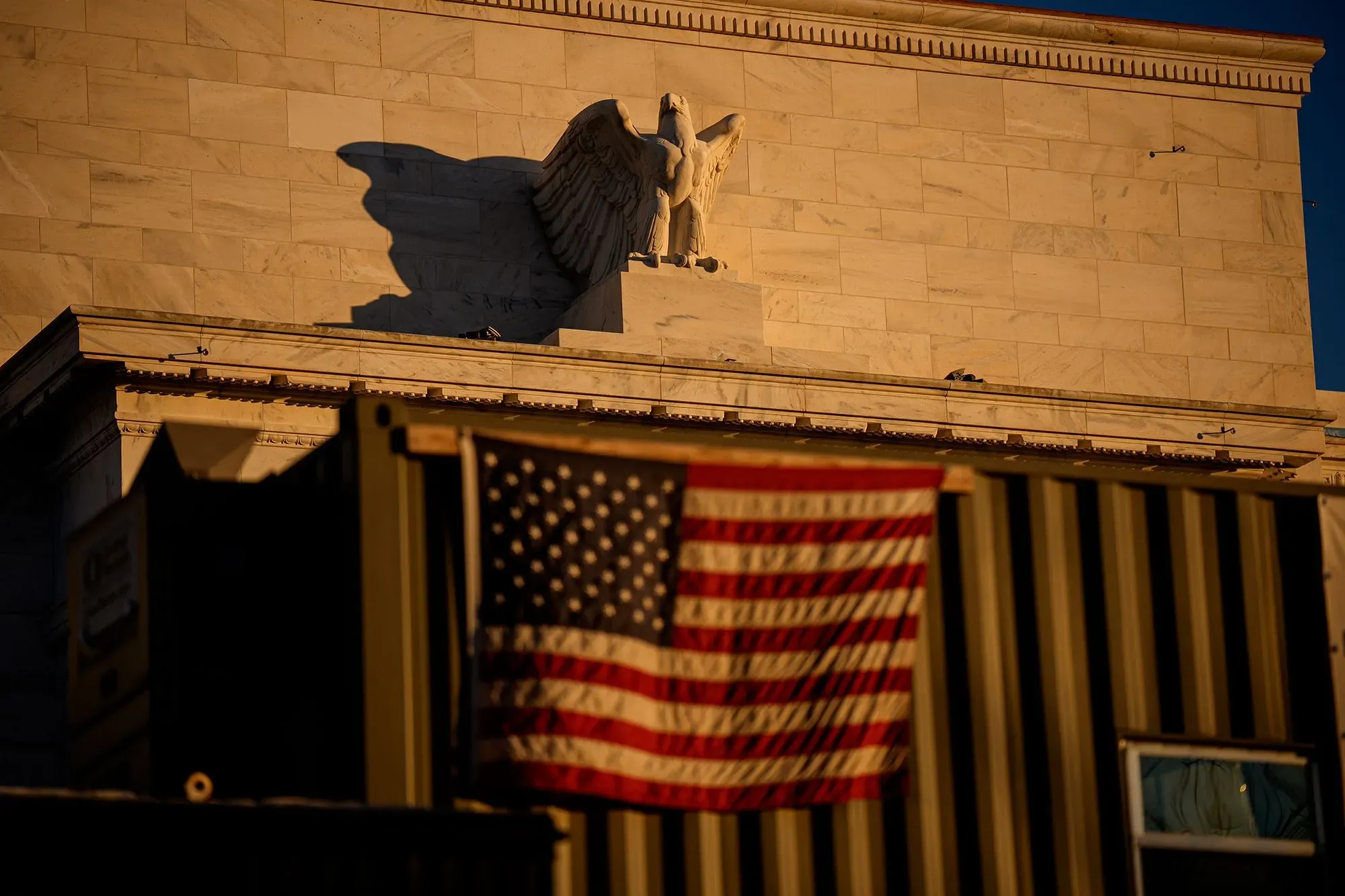 واجهة مبنى الاحتياطي الفيدرالي مع تمثال نسر، بينما ترفرف علم الولايات المتحدة في المقدمة، مما يعكس التوترات السياسية والاقتصادية الحالية.