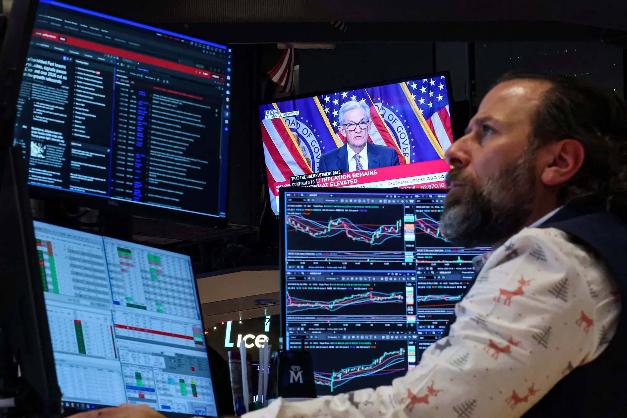 تاجر يتابع أسعار الأسهم والبيانات الاقتصادية على شاشات متعددة، بينما يظهر جيروم باول يتحدث في الخلفية، مما يعكس التوترات في سوق المال.