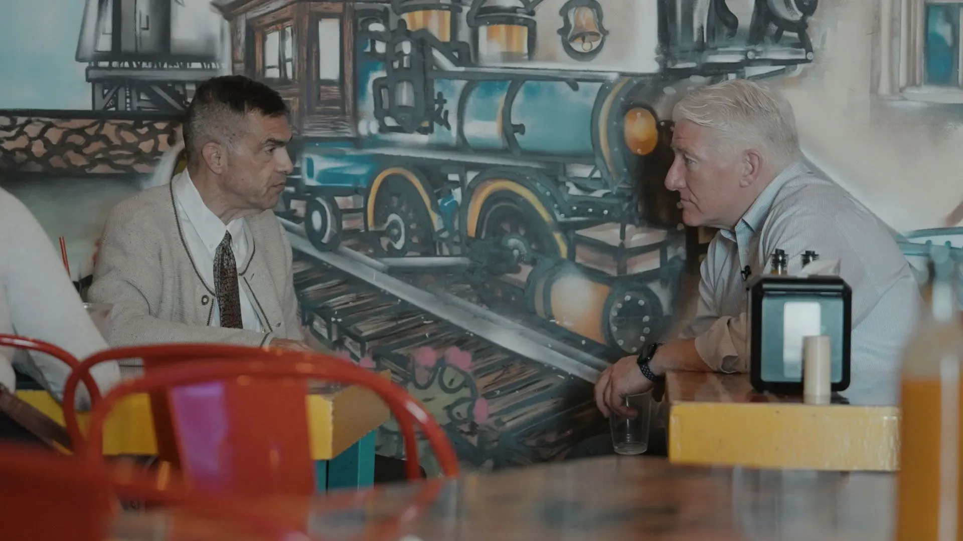 محادثة بين رجلين في مطعم ملون، مع خلفية تحتوي على رسومات لقطار، تعكس القضايا السياسية والاجتماعية في الانتخابات النصفية.