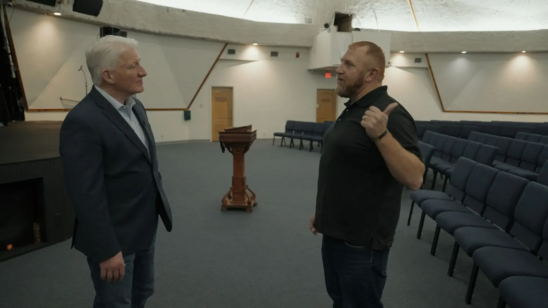 رجلان يتحدثان في قاعة فارغة، مع مقاعد مرتبة في صفوف، تعكس أجواء النقاش حول القضايا الانتخابية في تكساس.