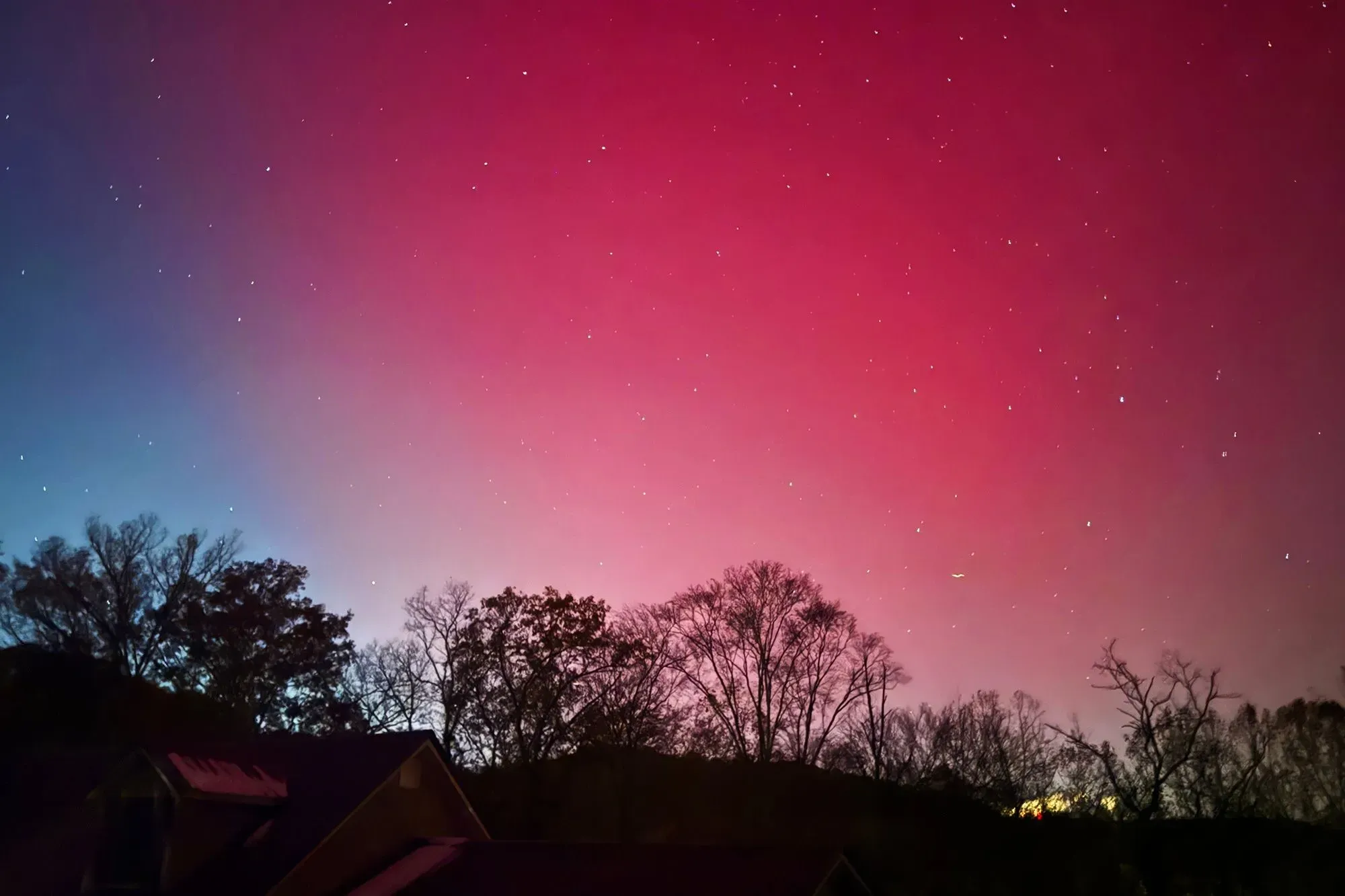 شفق ملون يظهر في السماء ليلاً، مع تأثيرات العواصف الشمسية، يضيء الأفق فوق الأشجار والمنازل.