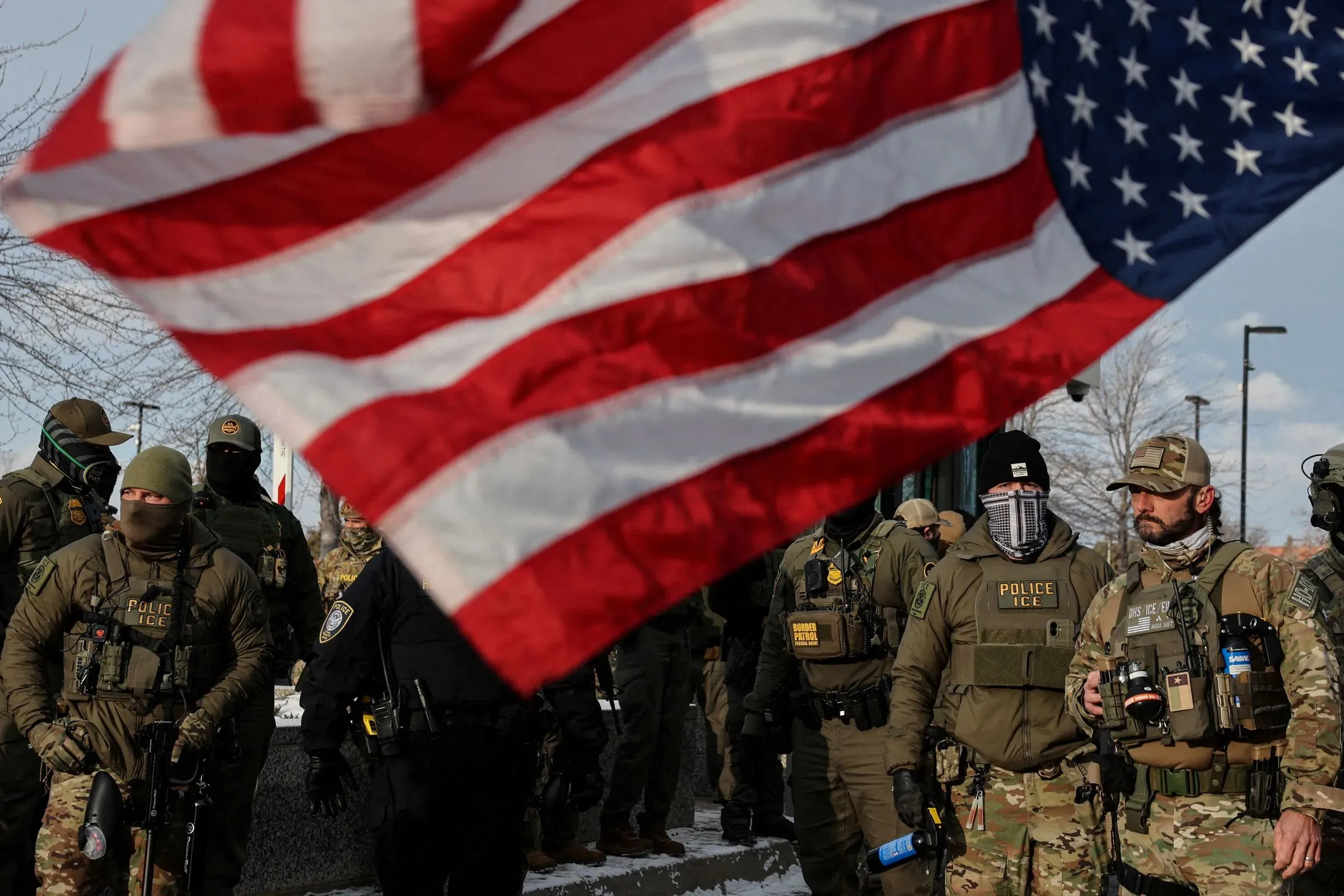 تظهر الصورة مجموعة من ضباط إنفاذ قوانين الهجرة والجمارك في مينيابوليس، مع العلم الأمريكي يرفرف في المقدمة، مما يعكس التوترات حول قضايا الهجرة.