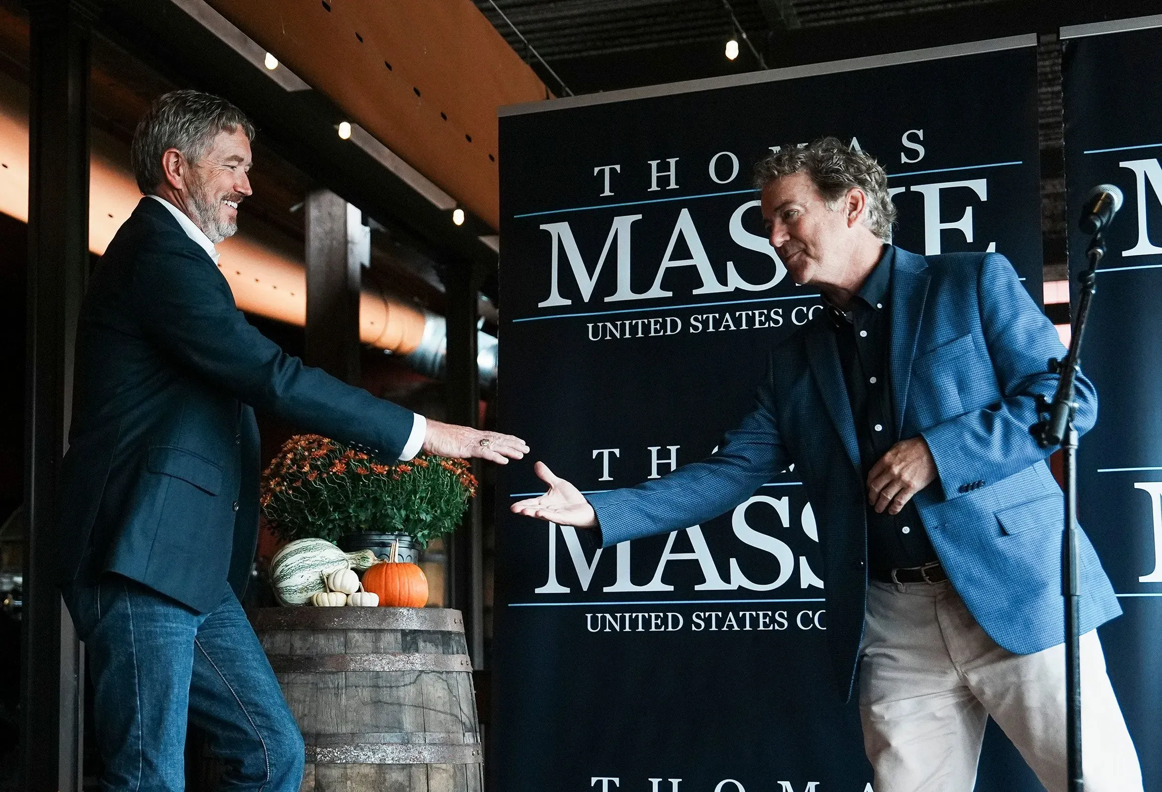 تظهر الصورة النائب توماس ماسي والسيناتور راند بول يتصافحان خلال حدث سياسي في كنتاكي، مع لافتة تدعم ماسي في خلفية الصورة.