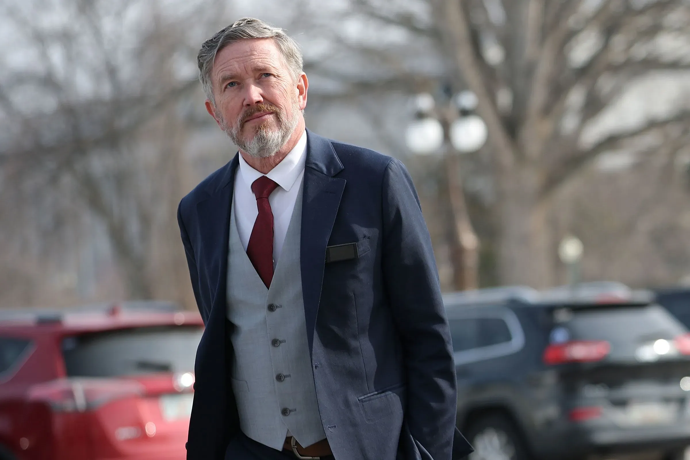 رجل يرتدي بدلة رسمية يسير في منطقة حضرية، وسط سيارات متوقفة، مع تعبير جاد على وجهه، في سياق سياسي متوتر في كنتاكي.