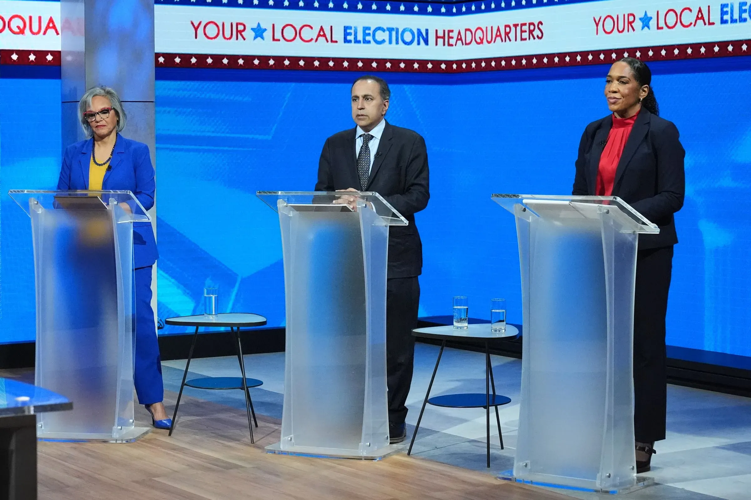 تظهر الصورة ثلاثة مرشحين في مناظرة انتخابية، حيث يتوسطهم طاولة شفافة، مع خلفية زرقاء تحمل شعار الانتخابات المحلية.