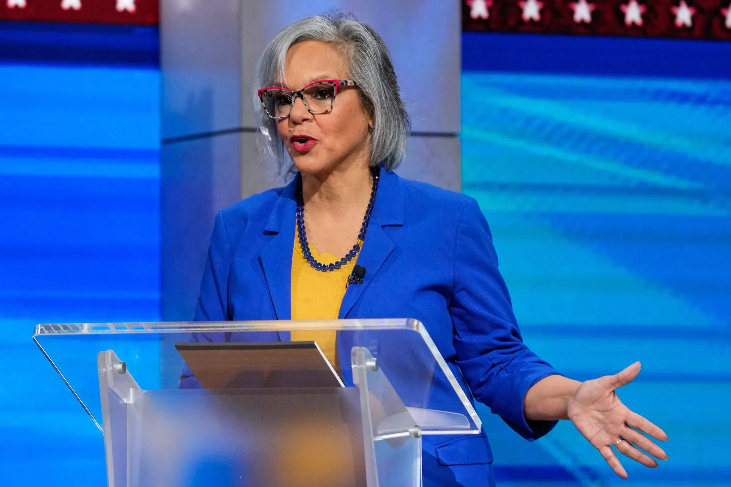 تظهر الصورة مرشحة ديمقراطية تتحدث في مناظرة انتخابية، مرتدية ملابس زرقاء، مع خلفية ملونة، تعكس التوترات السياسية في سباق إلينوي.