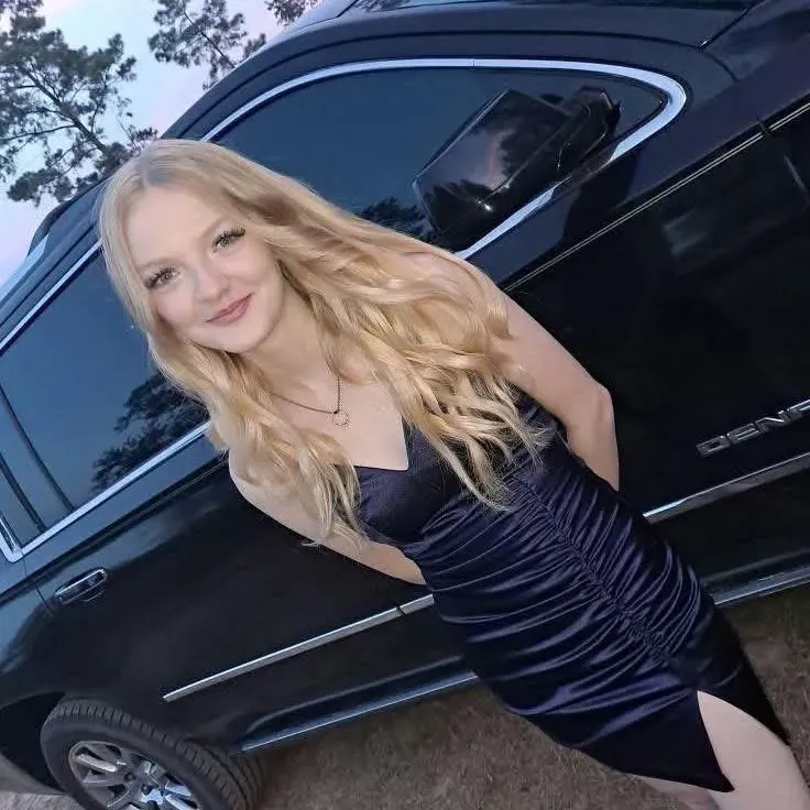 صورة لفتاة شابة ترتدي فستاناً أسود ضيقاً، تقف بجانب سيارة سوداء، مبتسمة وتظهر ملامح السعادة والثقة.