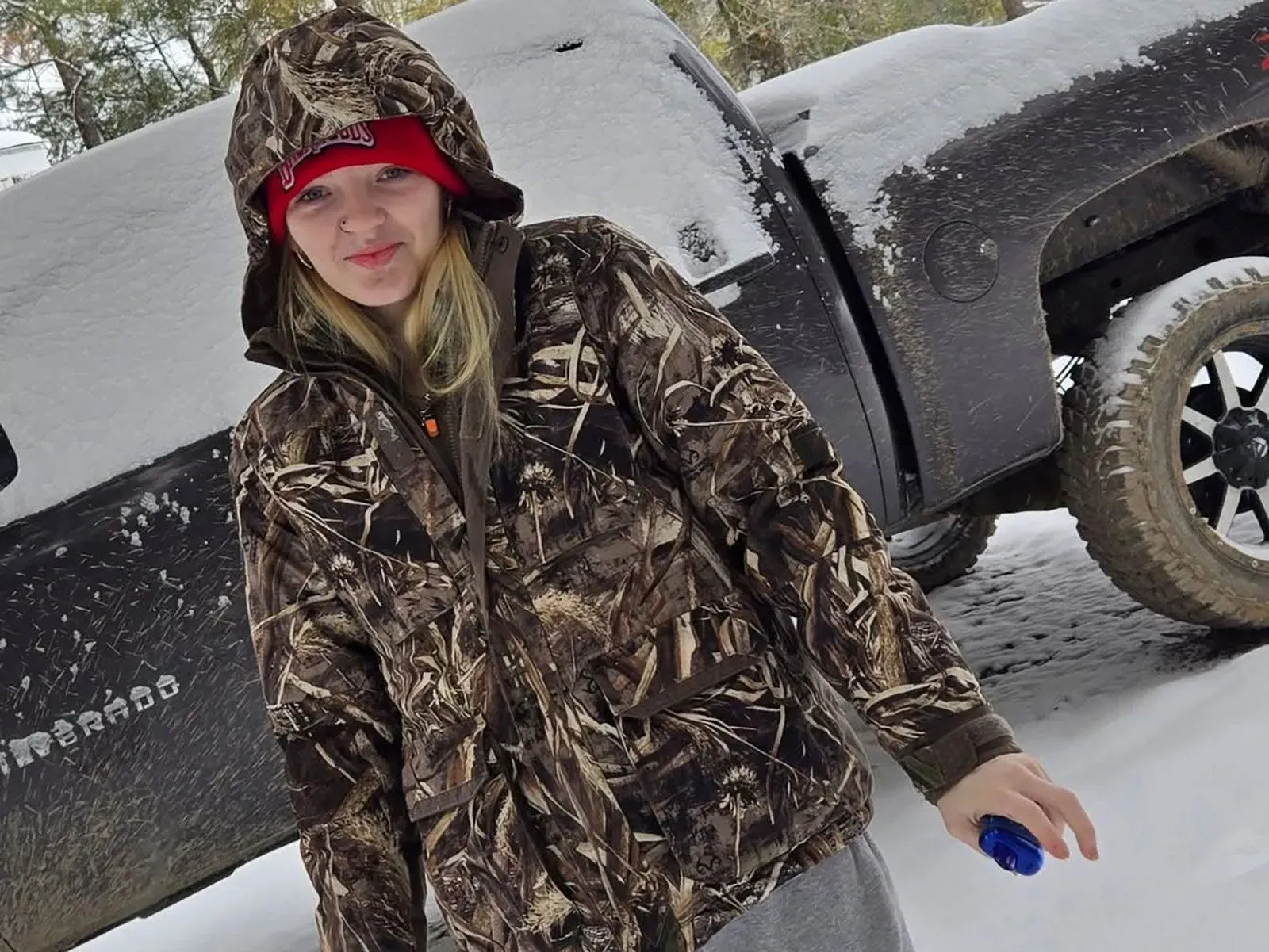 شابة ترتدي سترة مموهة وتبتسم في بيئة ثلجية، مع شاحنة في الخلفية. تعكس الصورة روح الأمل رغم الظروف الصعبة.