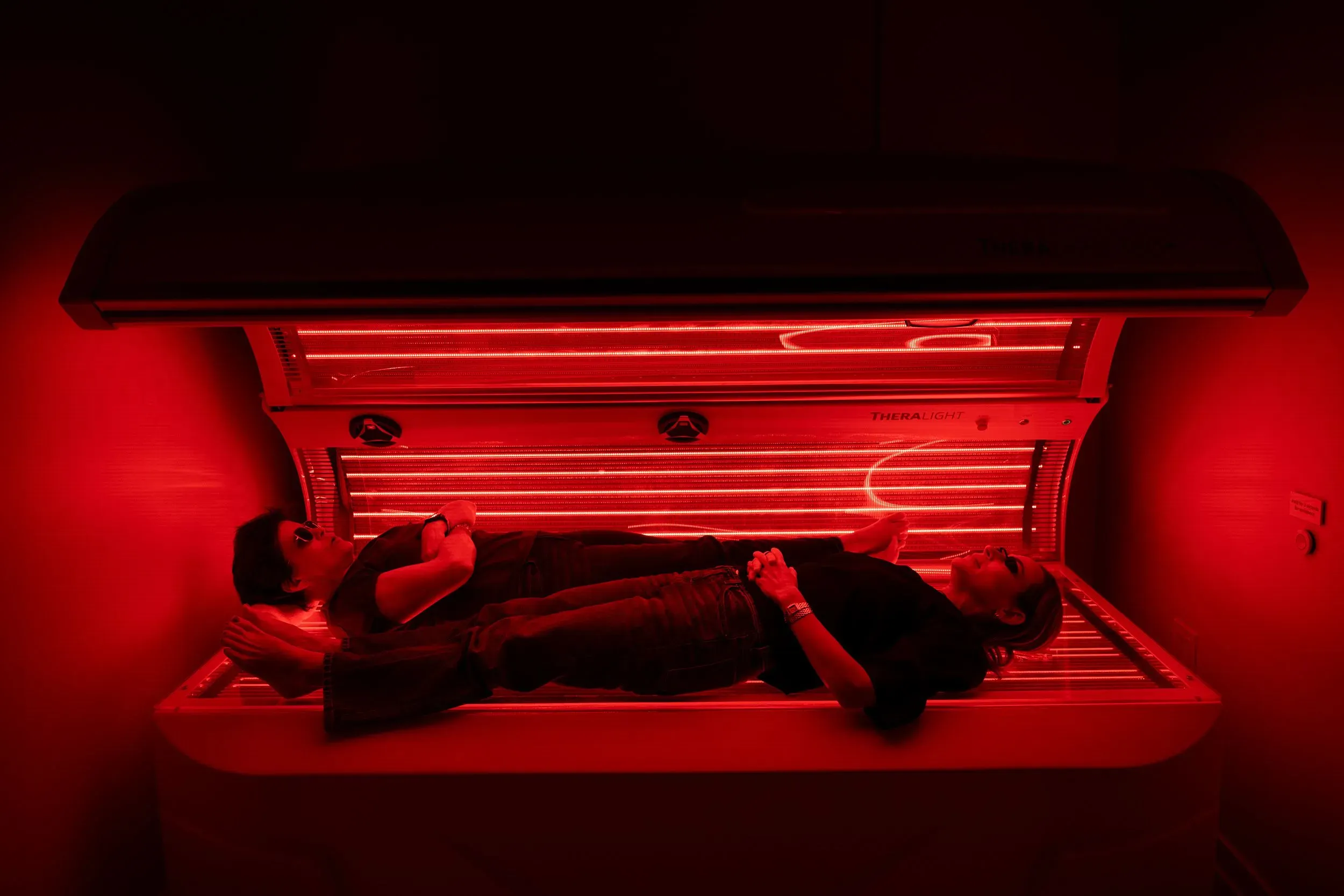 امرأتان تستلقيان تحت جهاز العلاج بالضوء الأحمر في غرفة مضاءة باللون الأحمر، مما يعكس استخدام هذه التقنية لتحسين صحة البشرة وتعزيز العافية.
