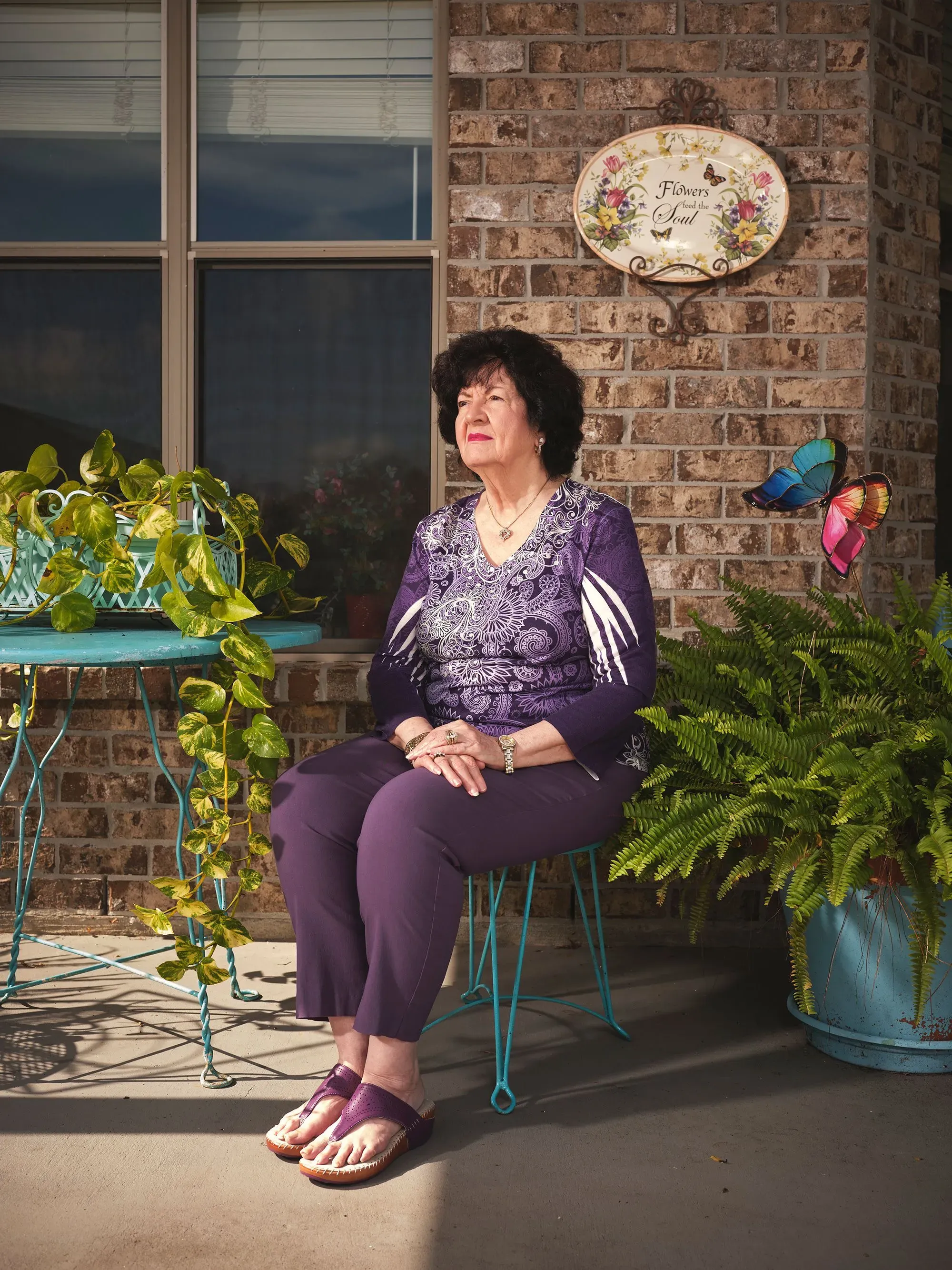 امرأة تجلس على كرسي في فناء منزلها، محاطة بالنباتات، وتظهر على الحائط لافتة مكتوب عليها "الزهور والروح"، تعكس روح المجتمع في بلاكيمين بلويزيانا.