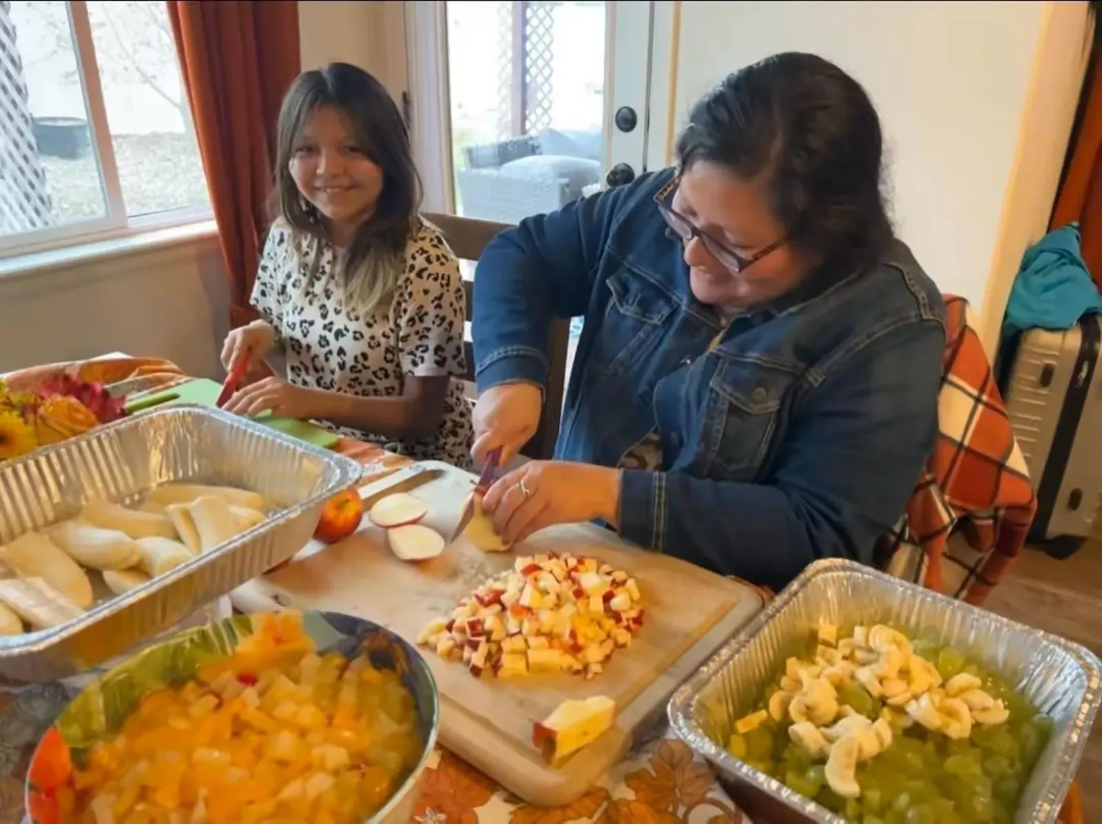 امرأة وفتاة تعملان معًا في المطبخ، حيث تقوم المرأة بتقطيع الفاكهة بينما تجلس الفتاة بجانبها مبتسمة، محاطة بأطباق مملوءة بالفواكه.