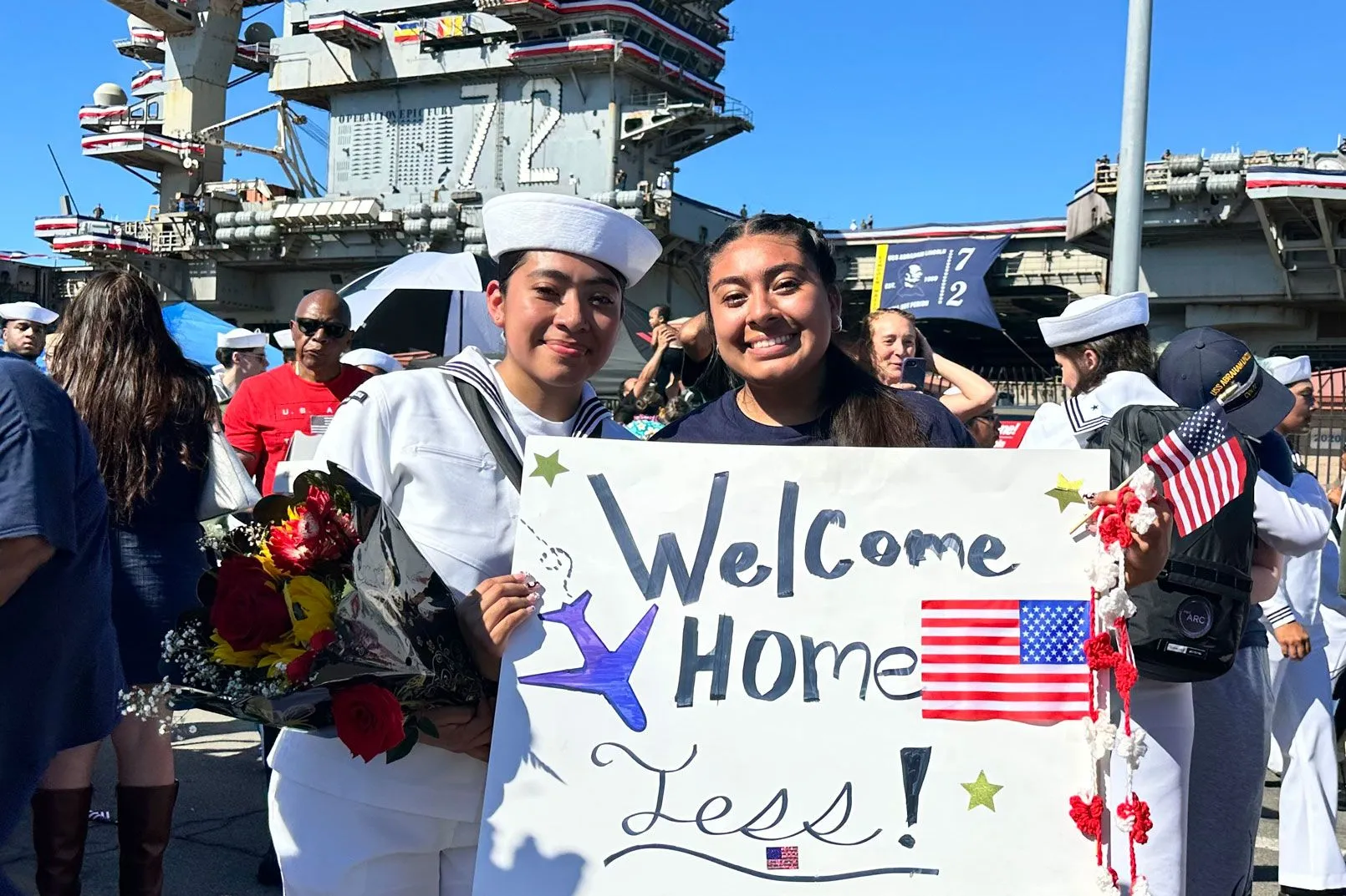 بحّارة أمريكية تحمل باقة زهور ولافتة ترحيب مكتوب عليها "Welcome Home Jess" خلال استقبال حاشد في ميناء سان دييغو بعد مهمة عسكرية طويلة.