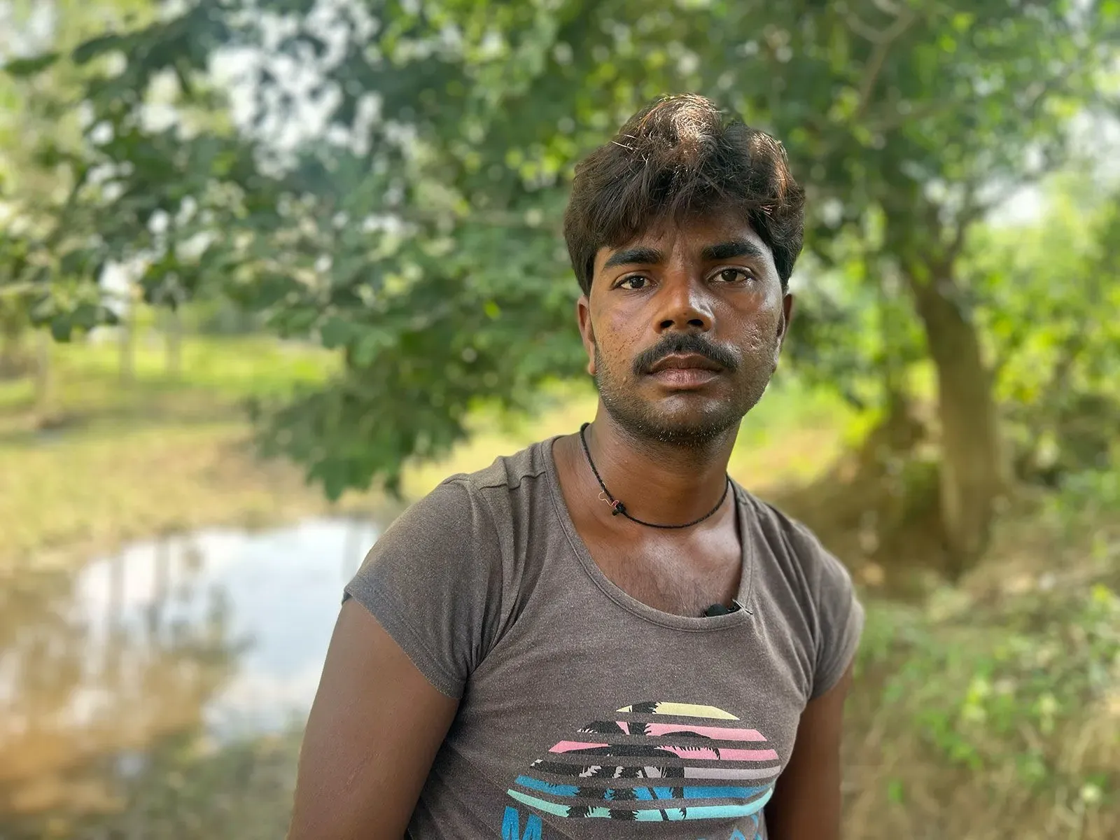 شاب يقف في منطقة ريفية شمال الهند، محاط بالأشجار والمياه. تعكس ملامحه القلق بعد اكتشافه طفلة حديثة الولادة مدفونة.