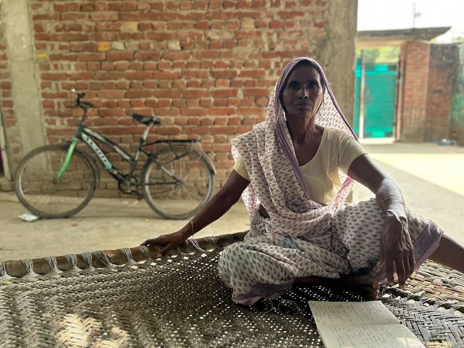 امرأة تجلس على سرير تقليدي في قرية هندية، ترتدي ساريًا، وبجانبها دراجة. تعكس ملامح وجهها القلق والتفكير العميق.