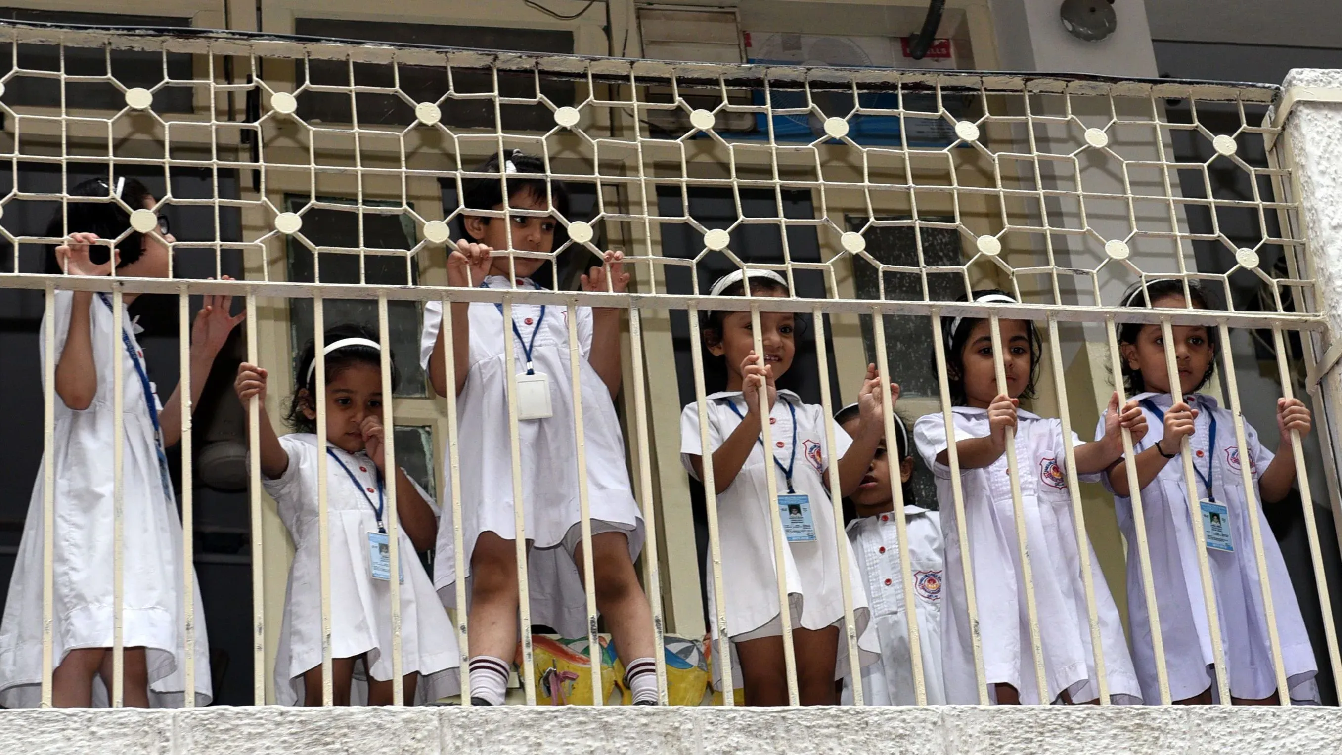 أطفال يرتدون زي المدرسة يقفون خلف سياج، يراقبون ما يحدث في الخارج، مما يعكس براءتهم وفضولهم.