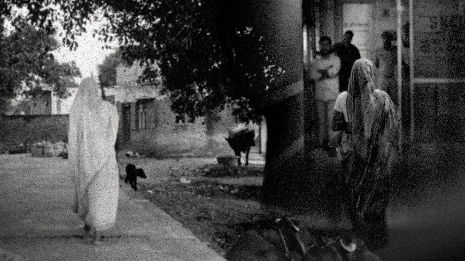 امرأة ترتدي ساريًا أبيض تمشي في شارع ريفي، بينما تظهر في الخلفية مجموعة من الأشخاص داخل مركز طبي، مما يعكس أجواء القرية في شمال الهند.