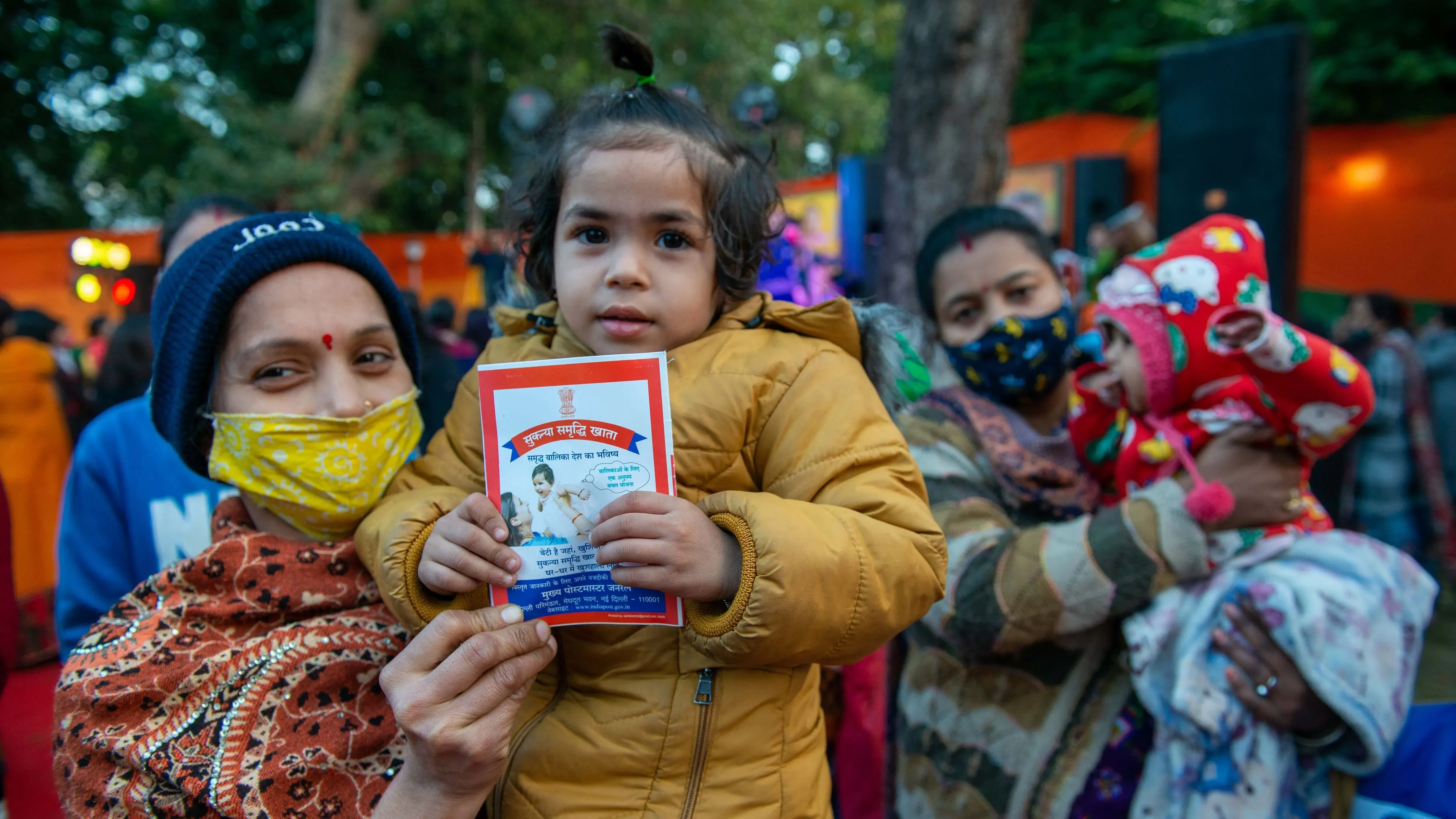 امرأة ترتدي وشاحًا ملونًا تحمل طفلة صغيرة ترتدي سترة صفراء، بينما تحمل امرأة أخرى طفلاً في حضنها. يظهر في الخلفية حشد من الناس في حدث عام.