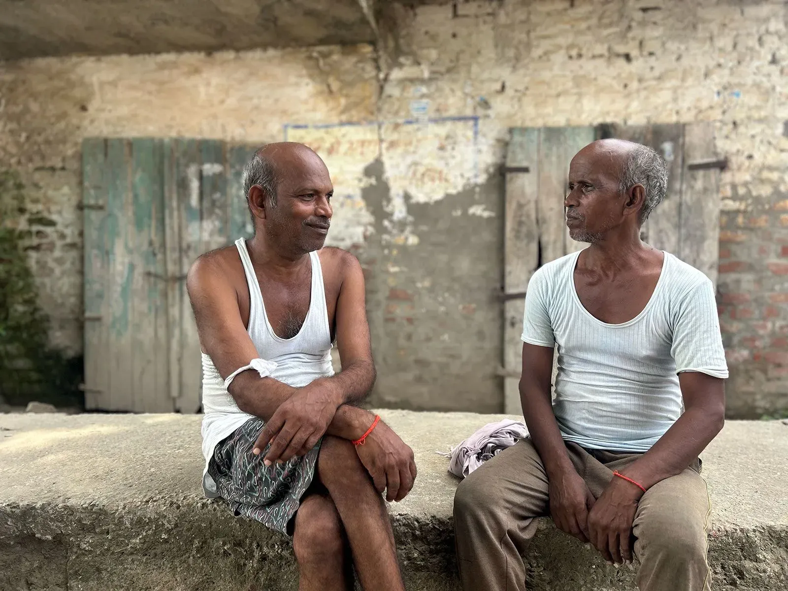 رجلان يجلسان على حافة جدار في قرية شمال الهند، يتحدثان معًا في جو غير رسمي، مع خلفية من الجدران الطينية والأخشاب.