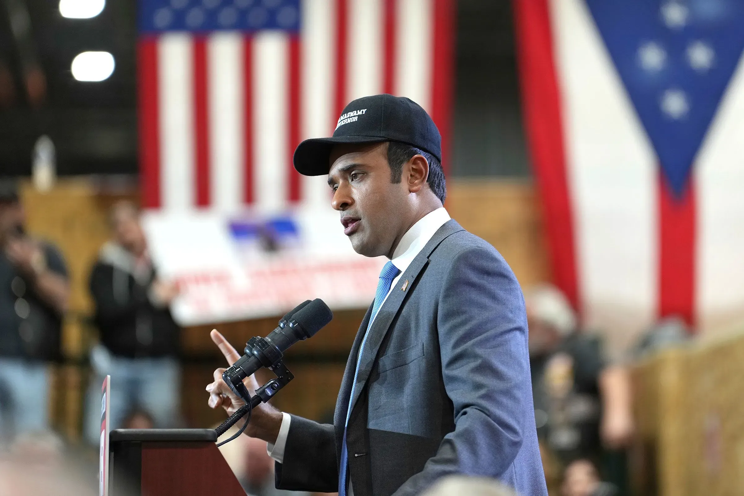 رجل يتحدث إلى حشد أثناء حدث سياسي، يرتدي بدلة وقبعة، مع أعلام أمريكية في الخلفية. يناقش قضايا الضرائب العقارية وتأثيرها على الأسر.