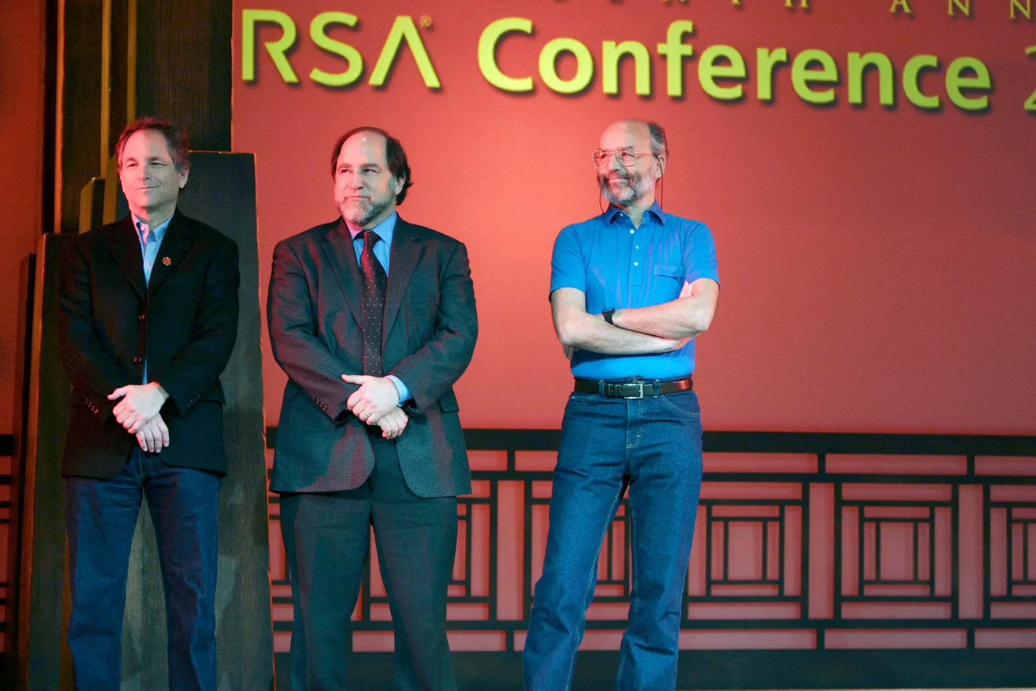 ثلاثة رجال يقفون على خشبة المسرح خلال مؤتمر RSA، مع خلفية حمراء تحمل شعار المؤتمر، يعكس أهمية الأمن الرقمي والحوسبة الكمومية.