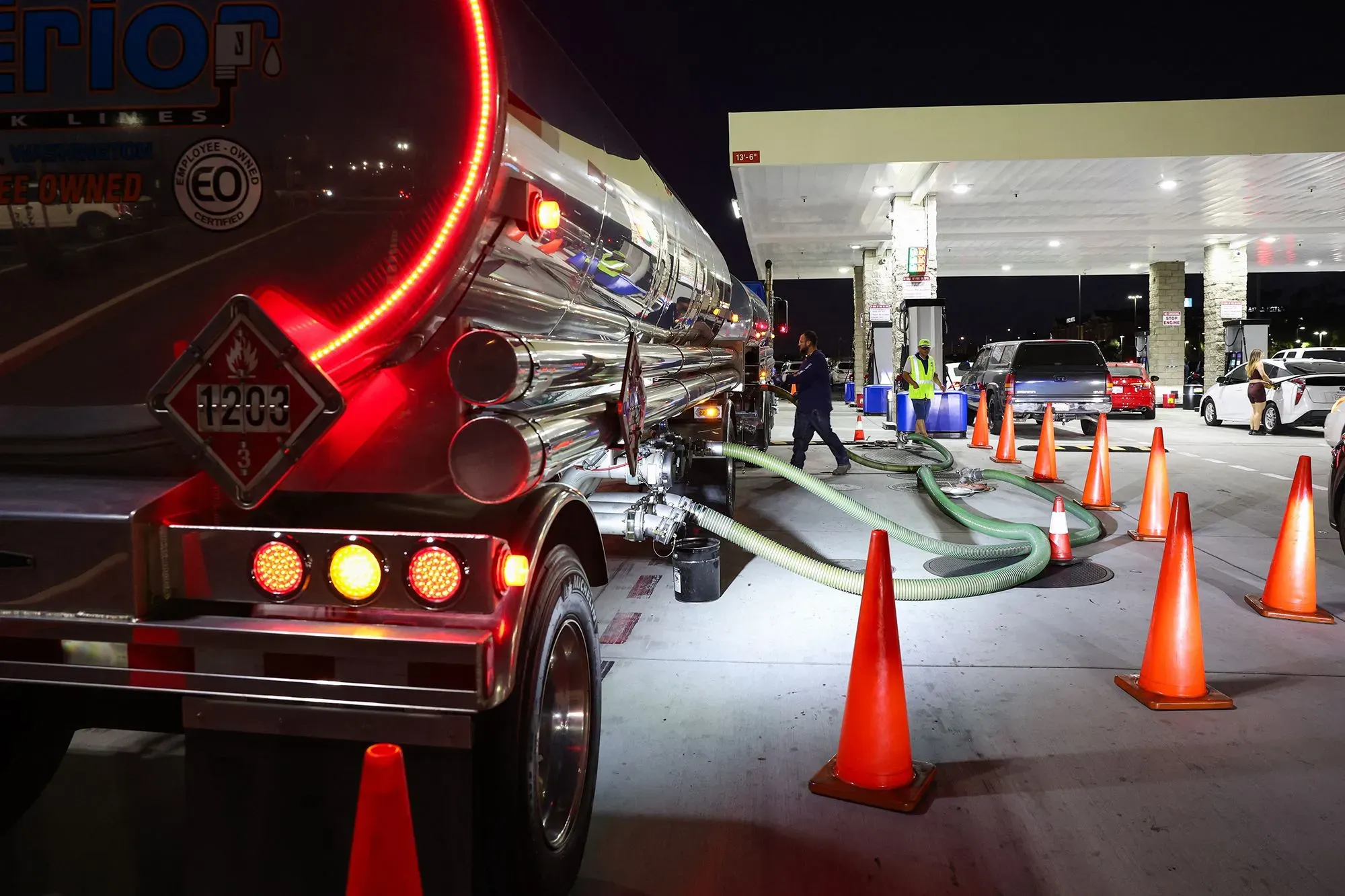 شاحنة وقود تقوم بتعبئة محطة بنزين في الليل، مع وجود حواجز مرورية وأشخاص يعملون، مما يعكس تأثير أسعار النفط على الاقتصاد الأمريكي.