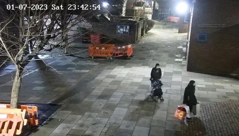 زوجان يسيران في الشارع ليلاً، حيث تحمل المرأة عربة أطفال بينما يحمل الرجل كيس بقالة. يوضح المشهد حالة القلق والتوتر المحيطة بهم.