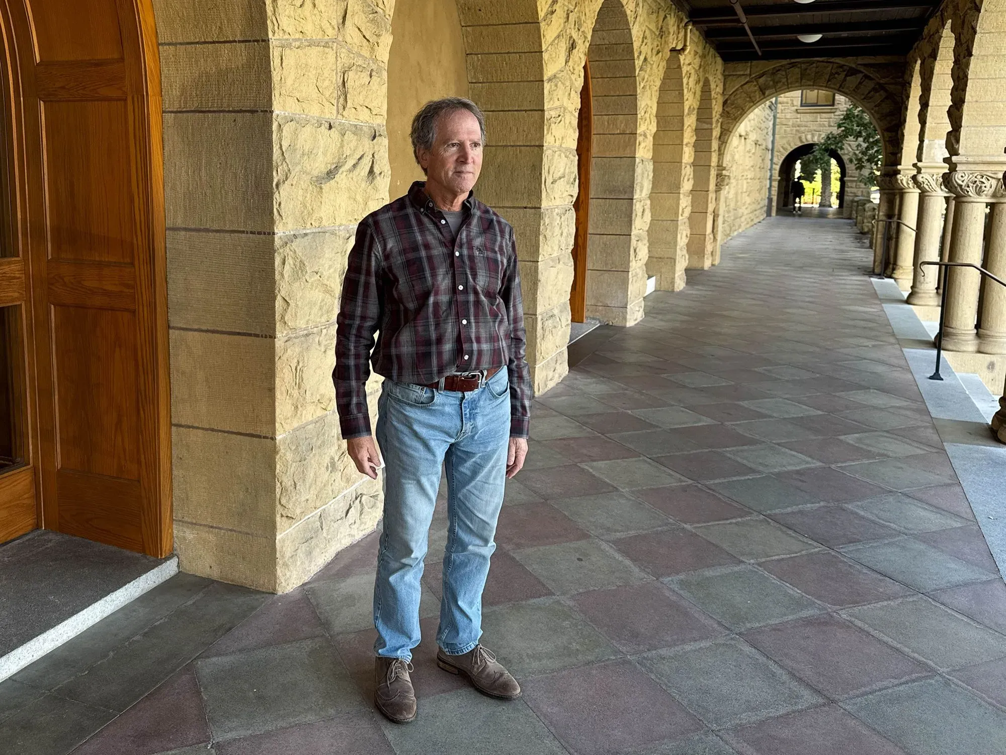 رجل يقف في ممر جامعة ستانفورد، يرتدي قميصًا مزخرفًا وبنطلون جينز، في خلفية معمارية تقليدية.