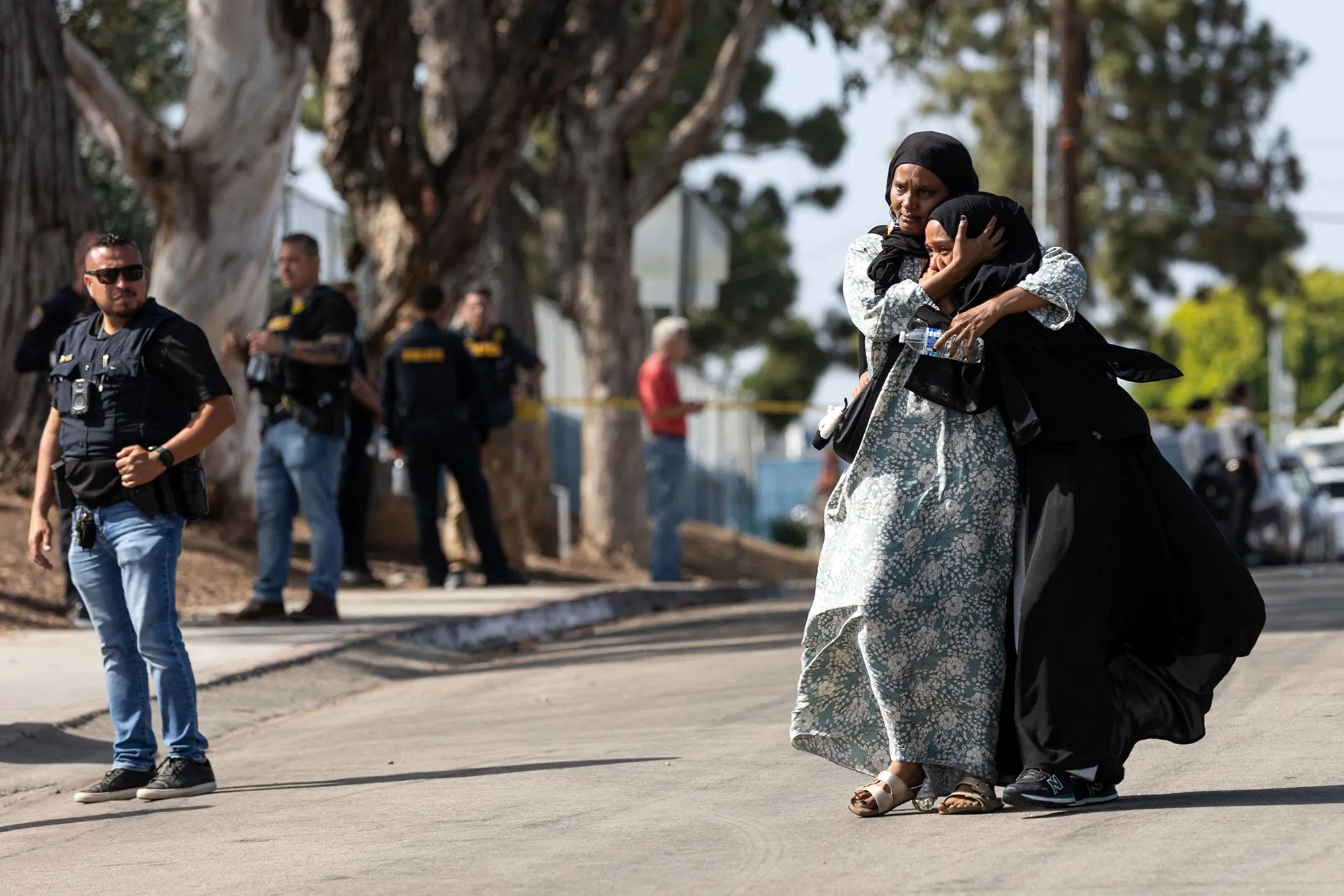 امرأتان ترتديان الحجاب، تتعانقان في حالة من الحزن، بينما يظهر رجال الشرطة في الخلفية بعد حادث إطلاق نار في مركز إسلامي في سان دييغو.