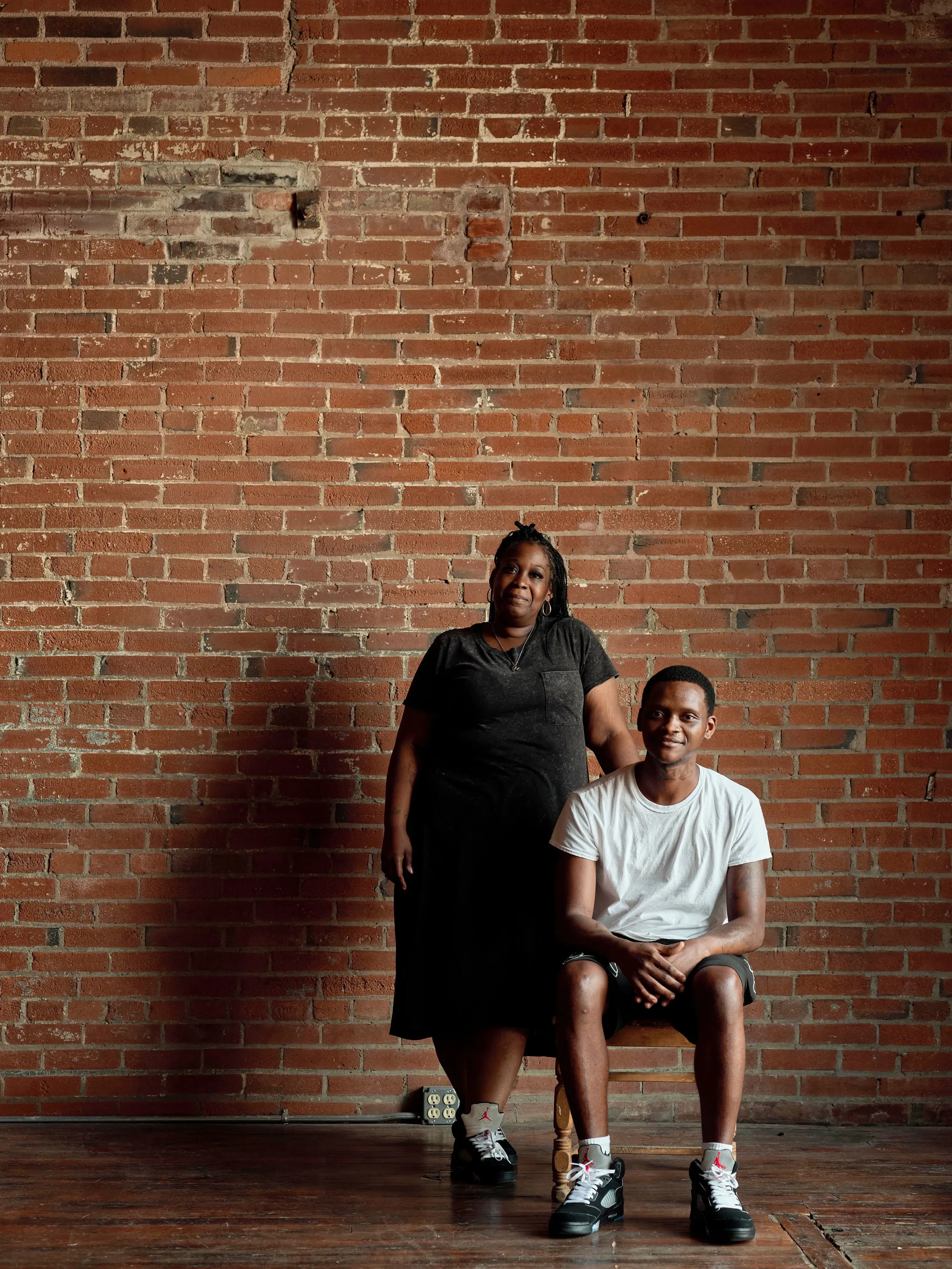 امرأة ترتدي ملابس سوداء تقف خلف شاب يجلس على كرسي، في غرفة ذات جدران من الطوب، تعكس قصة حياة وتجارب معقدة.