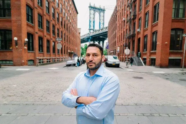 رجل يقف بثقة في شارع ضيق بين مباني من الطوب الأحمر، مع جسر مانهاتن في الخلفية، يعكس موضوعات الحياة والتحديات.