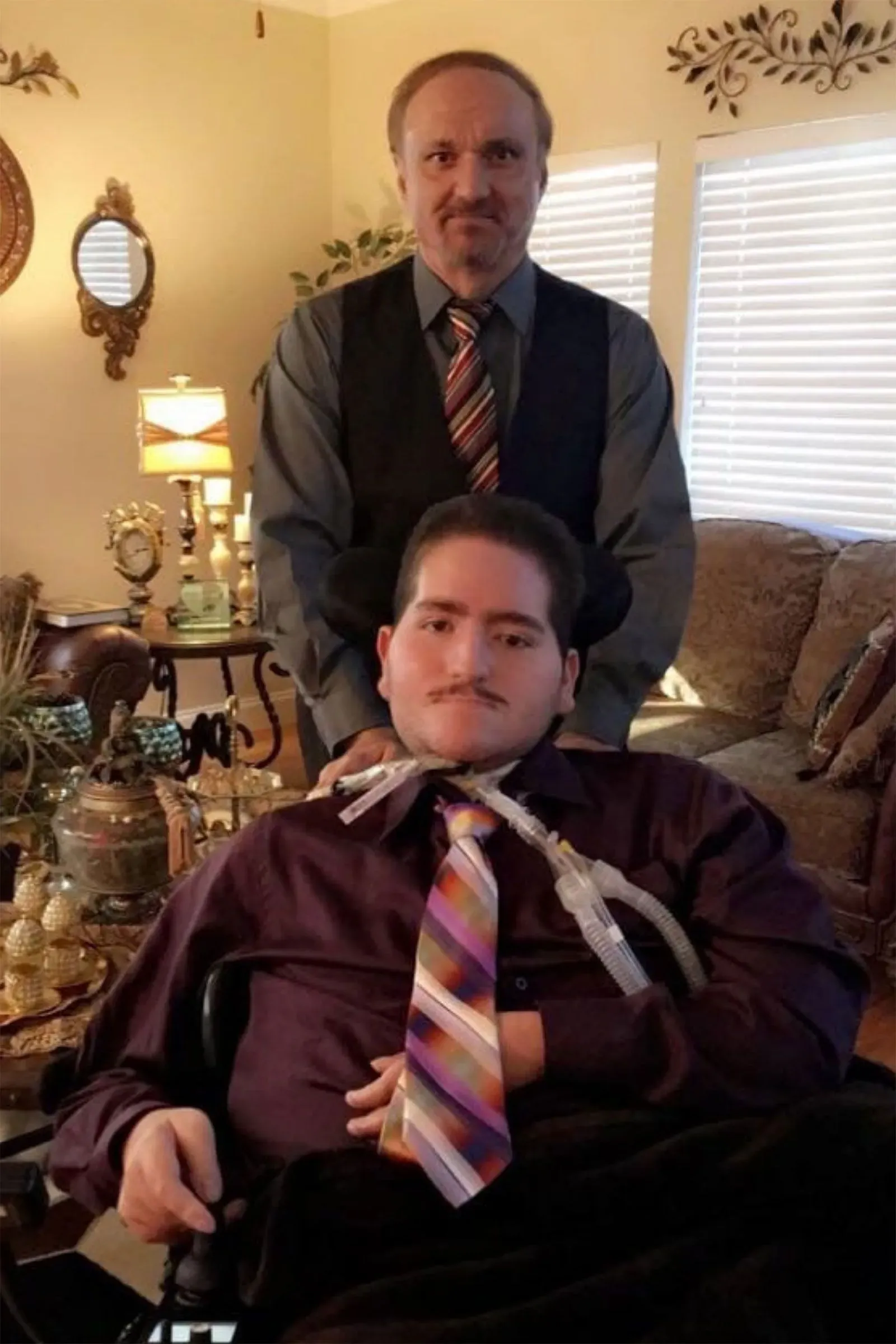 صورة تُظهر وائل طرابيشي، شاب يعاني من مرض بومبي، يجلس على كرسي متحرك بينما يقف والده ماهر خلفه، في لحظة عائلية مؤثرة.