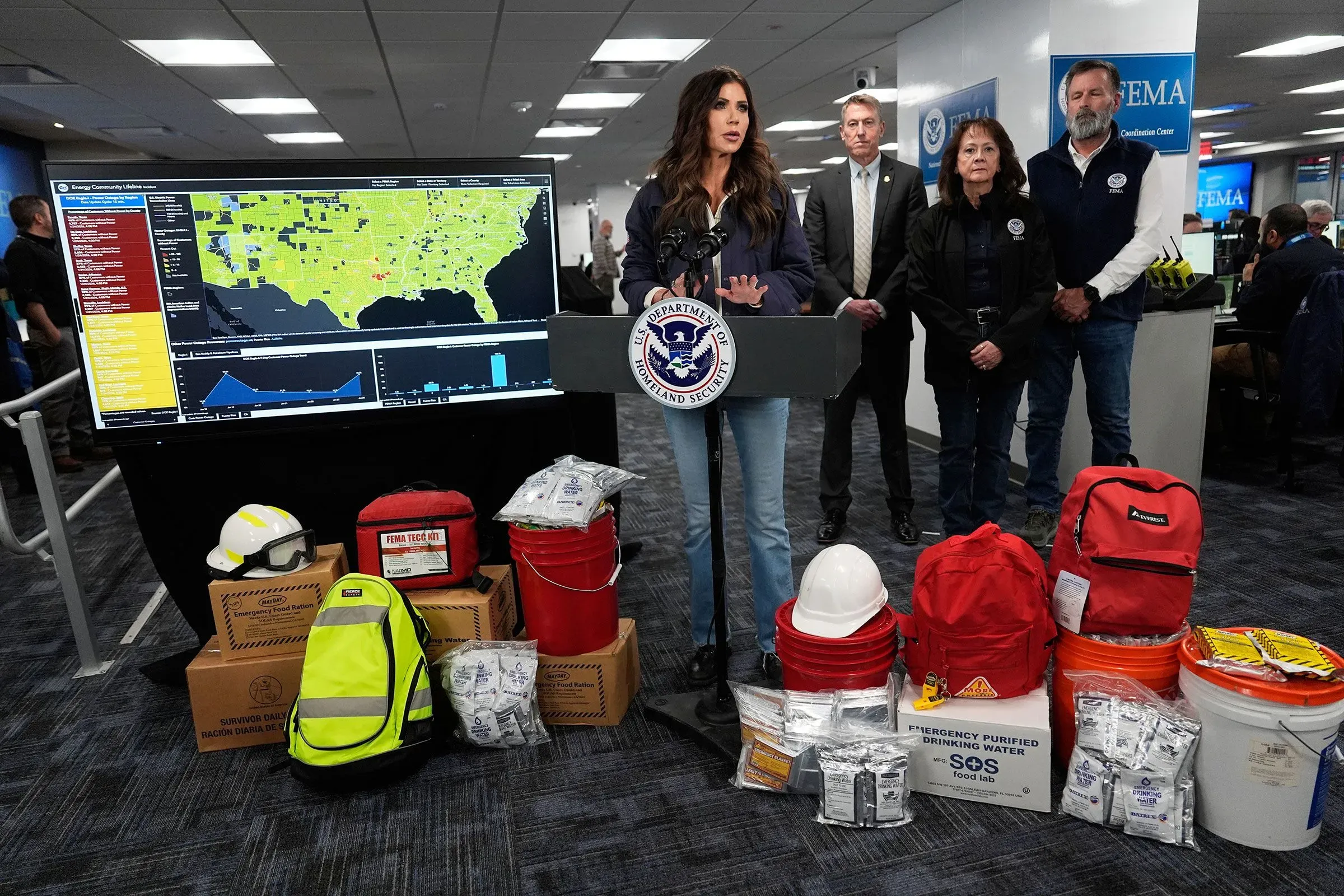 مسؤولة في وكالة إدارة الطوارئ الفيدرالية تتحدث أمام وسائل الإعلام، مع خريطة توضح مناطق العواصف وأدوات الطوارئ أمامها.