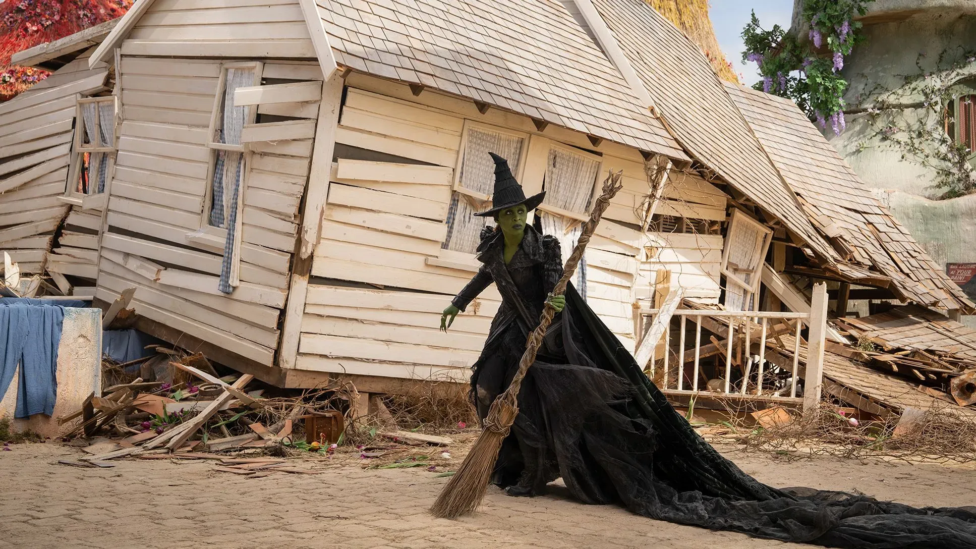 ساحرة ذات بشرة خضراء ترتدي رداءً أسود وقبعة مدببة، تحمل عصا مكنسة، وتقف أمام منزل مائل في مشهد يعكس قوة شخصيتها.