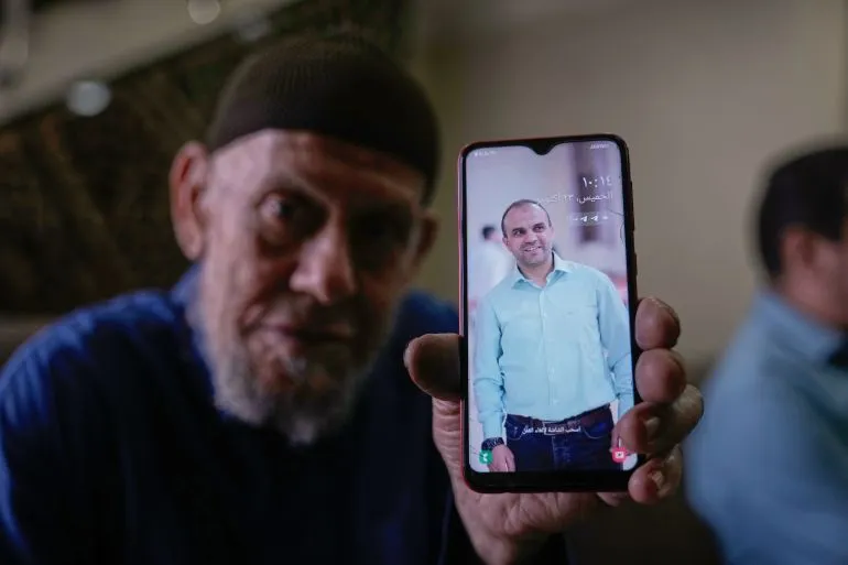 رجل مسن يحمل هاتفًا ذكيًا يظهر صورة لشاب مبتسم، تعبيرًا عن الحزن والذكريات في سياق القضايا الإنسانية في فلسطين.
