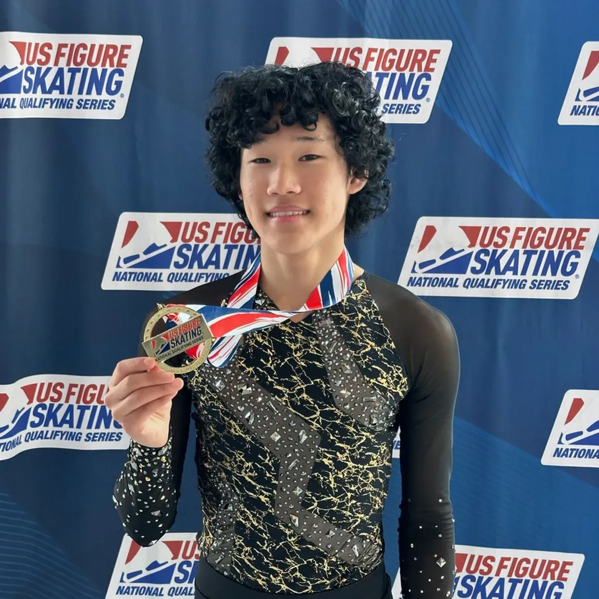 شاب يحمل ميدالية ذهبية في التزلج الفني على الجليد، مبتسمًا أمام خلفية تحمل شعار "US Figure Skating".