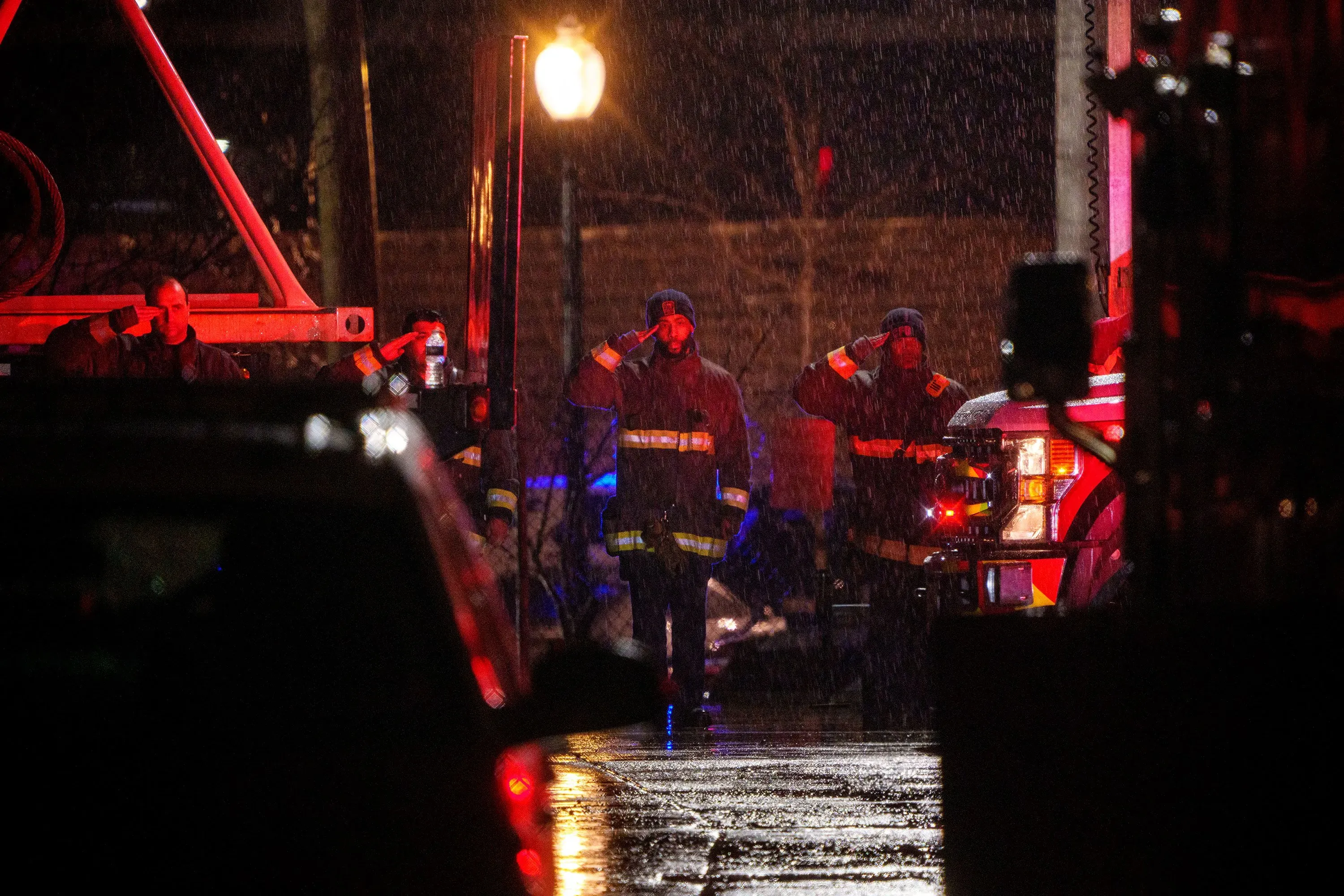 رجال الإطفاء والإنقاذ يقفون تحت المطر، يحيون تكريمًا لضحايا حادث تحطم الطائرة في بوتوماك، مع أضواء الطوارئ تضيء المكان.