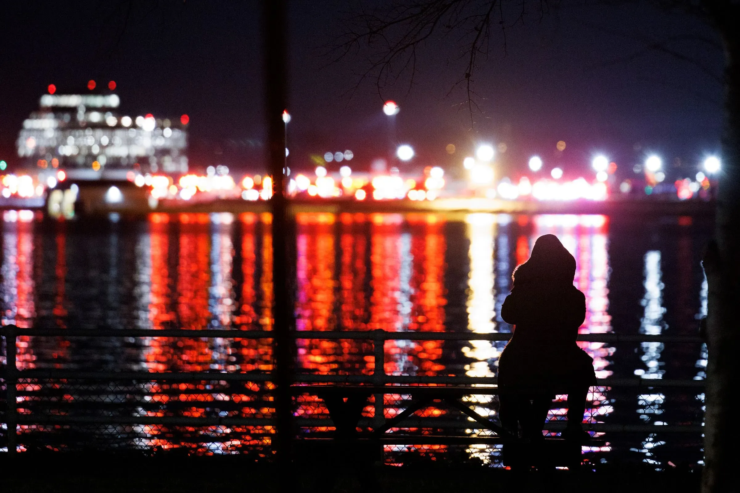 شخص يقف على ضفة نهر بوتوماك ليلاً، محاطاً بأضواء الطوارئ المتلألئة من سيارات الإسعاف والإطفاء، مما يعكس أجواء الحادث المأساوي.