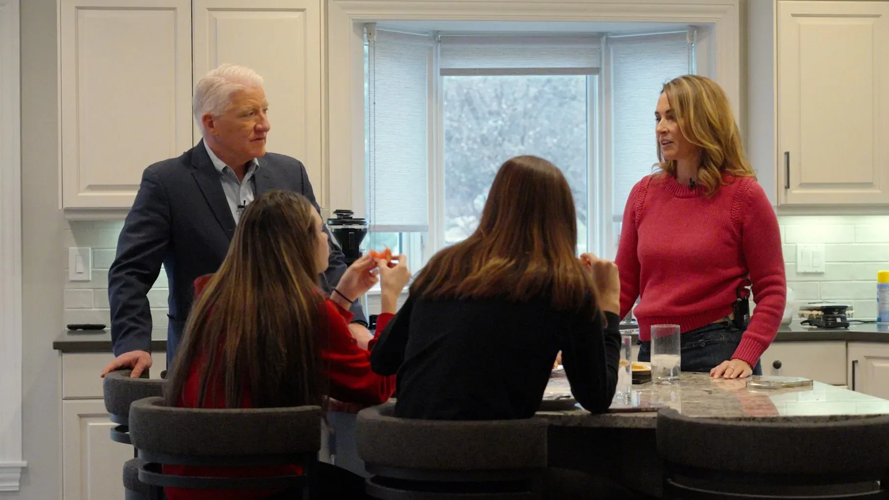 عائلة تتحدث حول طاولة في مطبخ حديث، حيث يظهر شخصان بالغين وفتاتان. تعكس الأجواء تفاعلًا عائليًا ودافئًا.