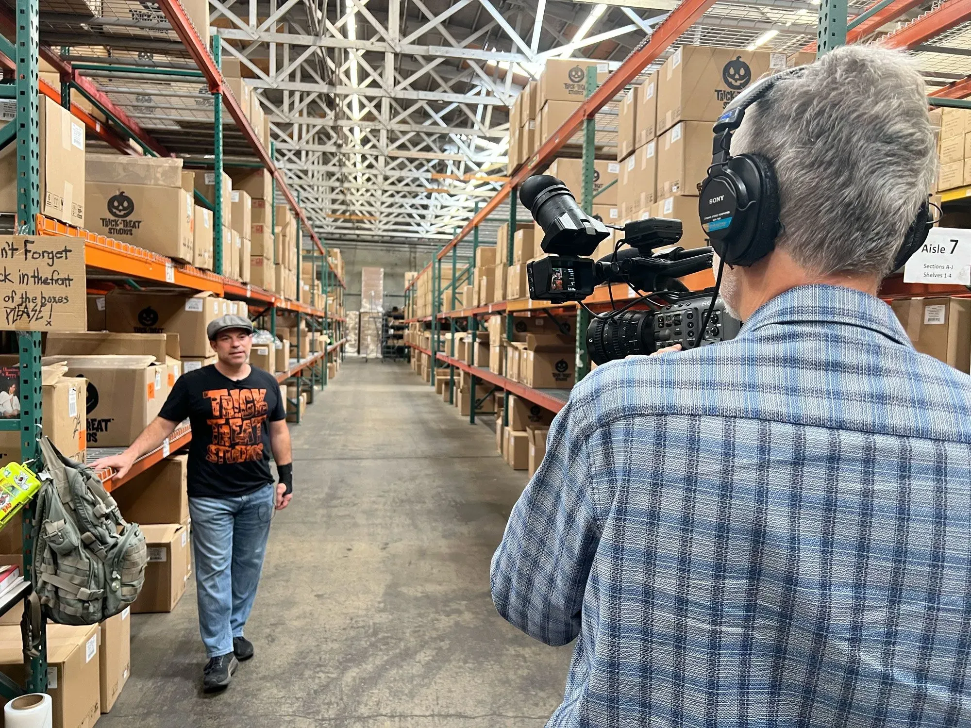 رجل يرتدي قميصًا أسود يحمل عبارة "Trick or Treat" يسير في مستودع مليء بالصناديق، بينما يقوم مصور بتوثيق المشهد.